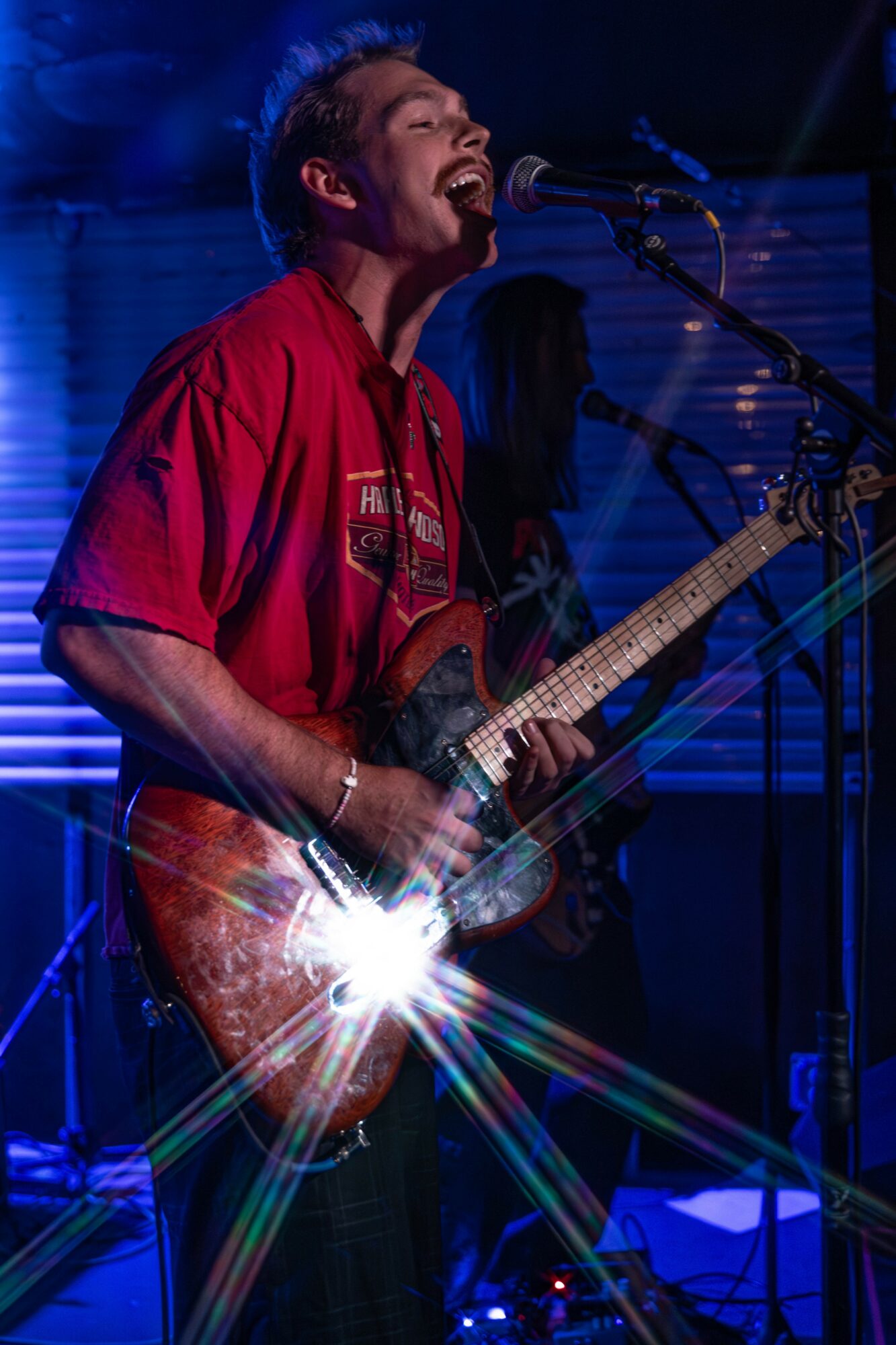 Person singing and playing guitar on stage with microphone, wearing red shirt, blue background, stage lighting, and musical equipment.