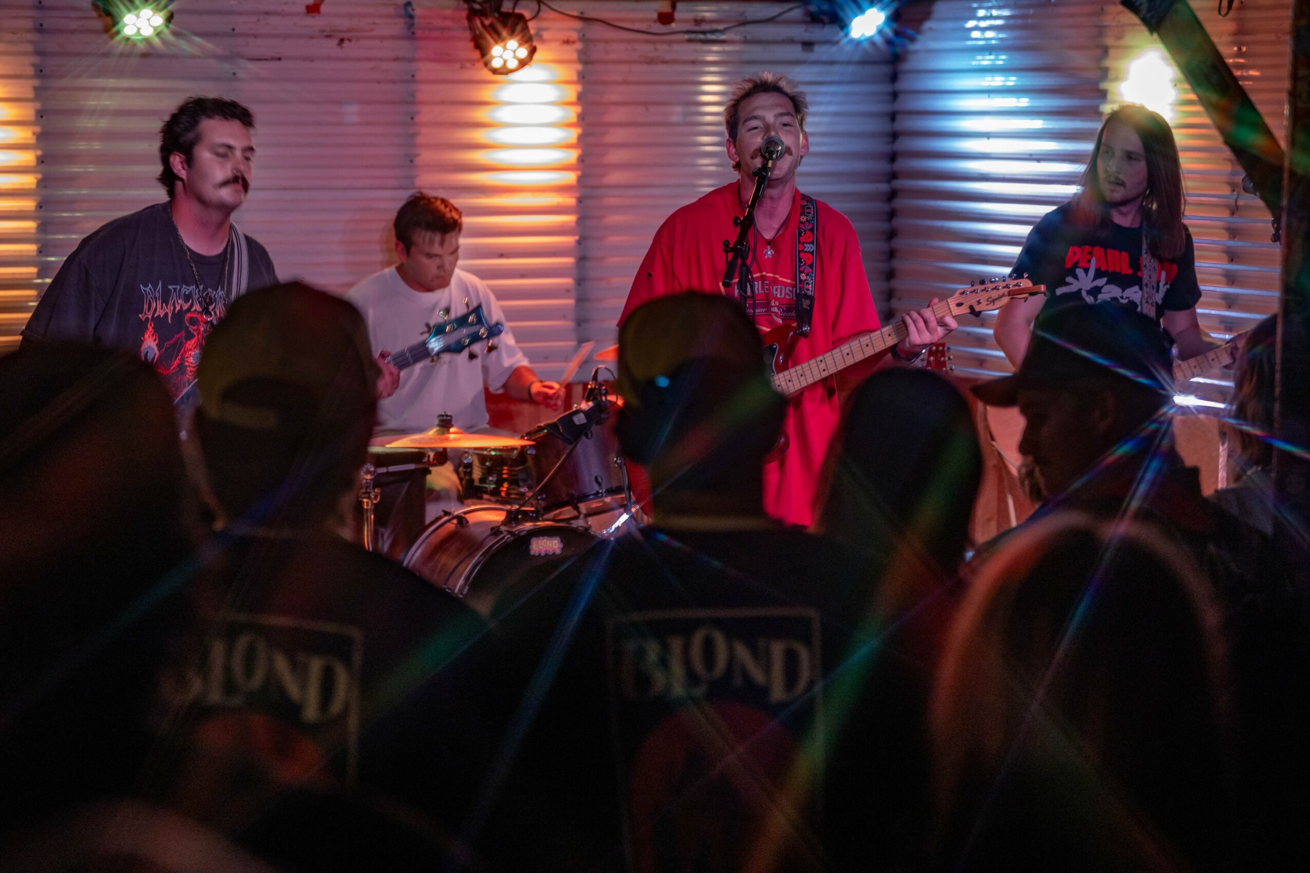 Band performs on stage with colorful lighting, audience in foreground, four musicians playing instruments, one singing, lively atmosphere.