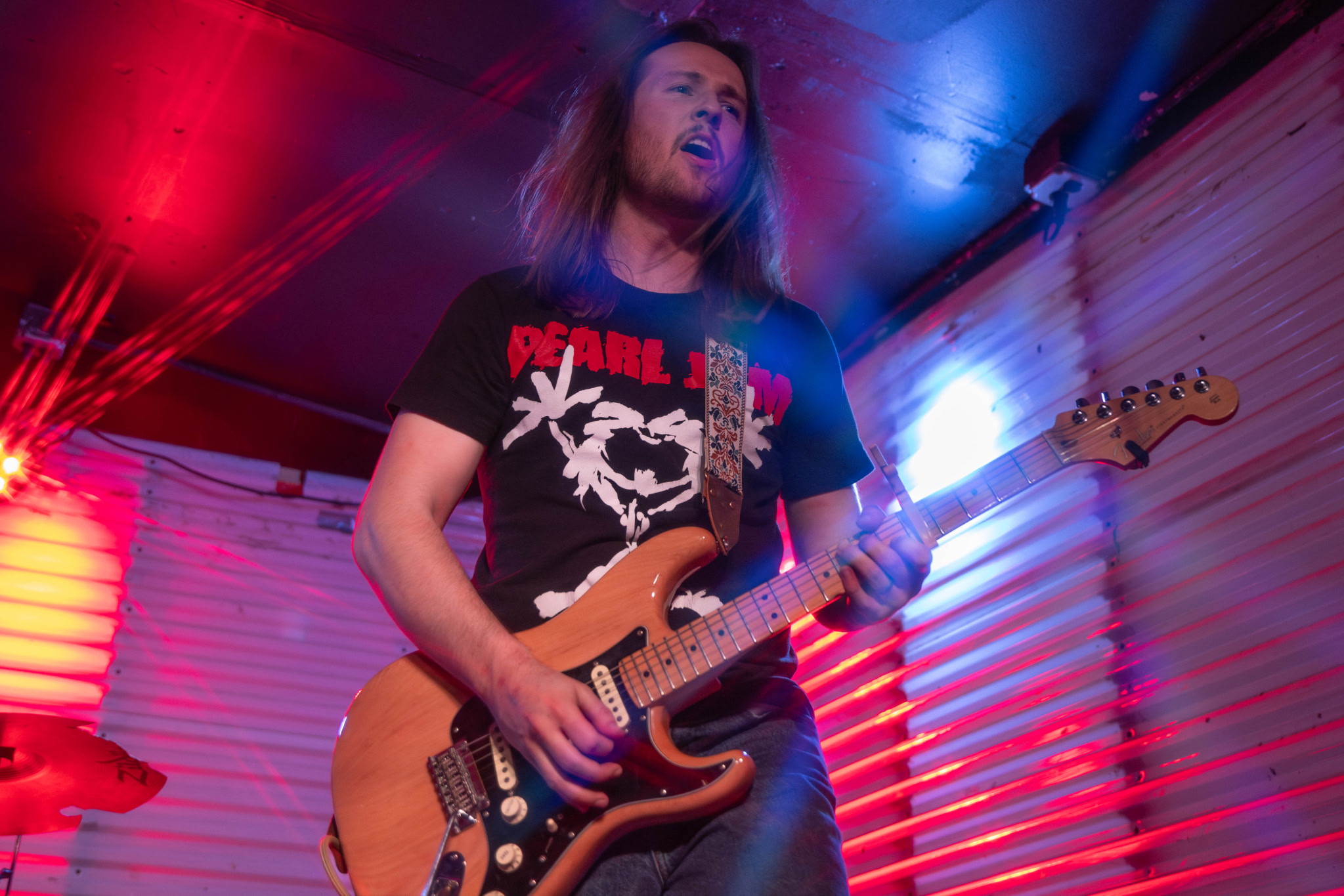 Person playing an electric guitar on stage with colorful lighting, wearing a black t-shirt with white text and graphics.