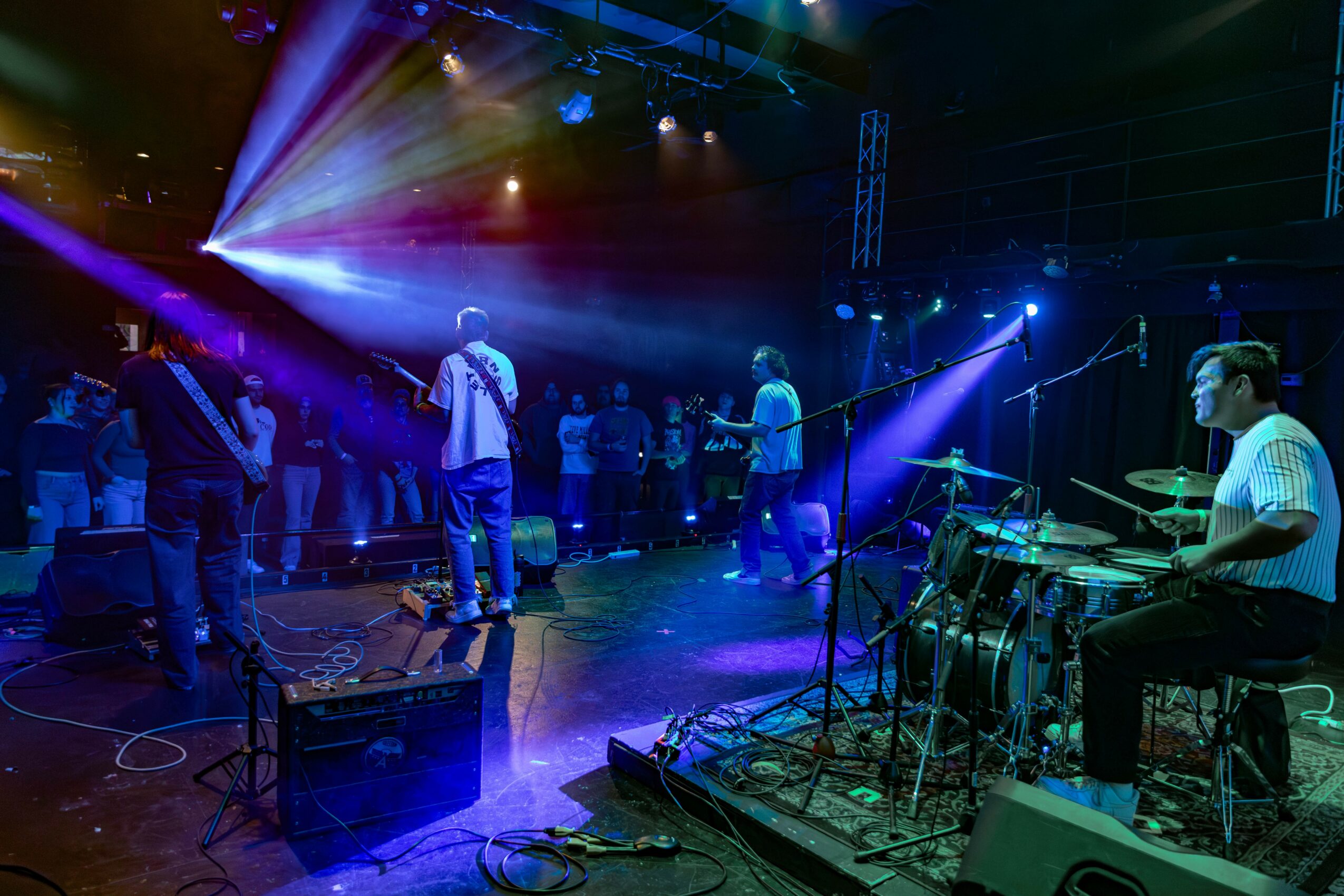 Band performs on stage with colorful lights, audience watching in a dark venue, drummer playing on the right, microphones set up.