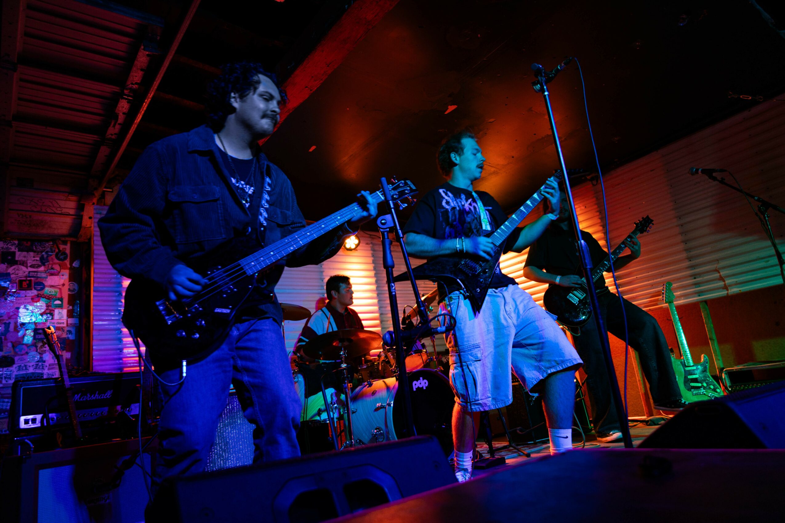 Band performing on stage with three musicians playing guitars and a drummer in the background, colorful stage lighting.