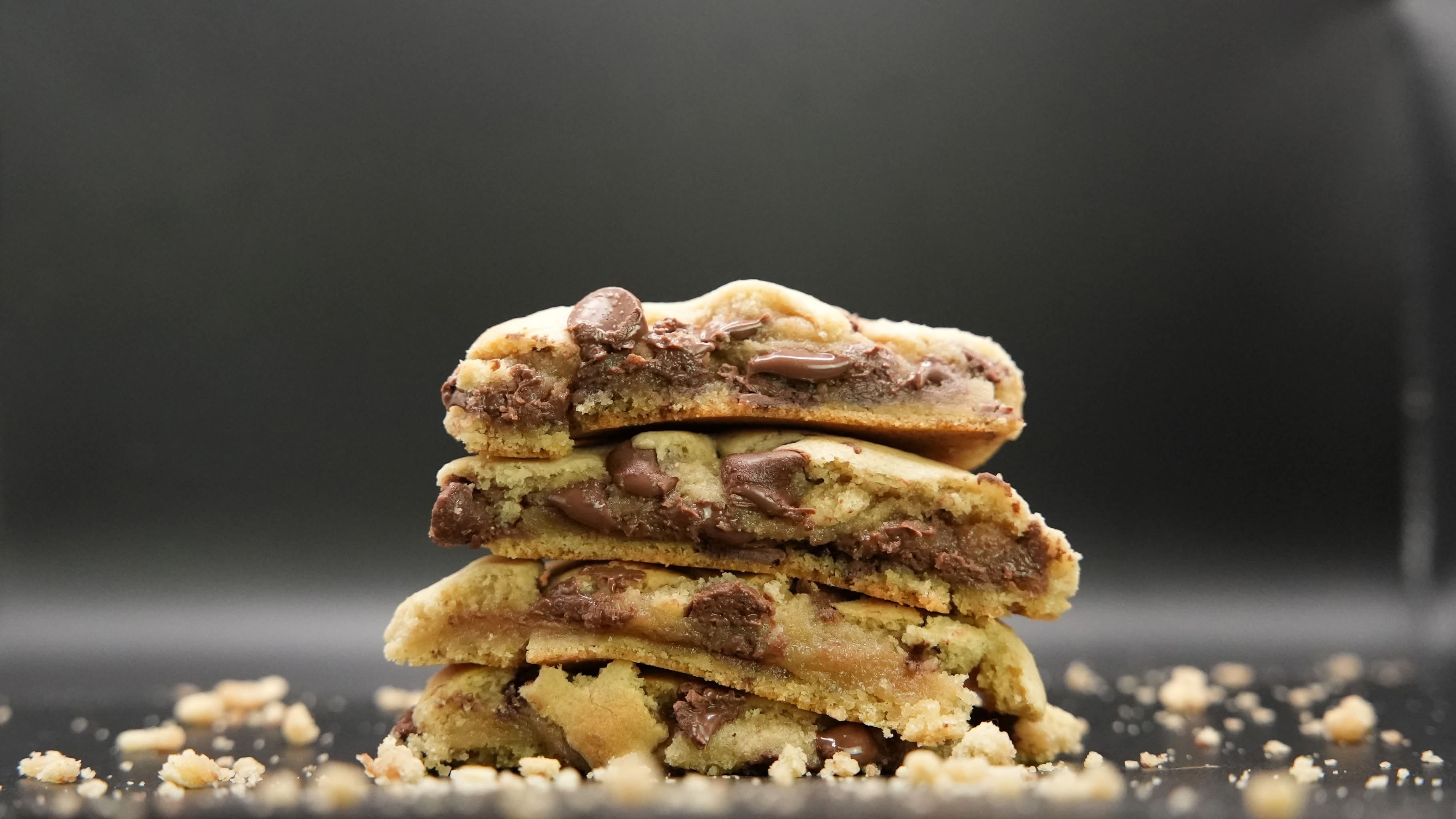 Stack of four chocolate chip cookies with crumbs scattered around, against a dark background.