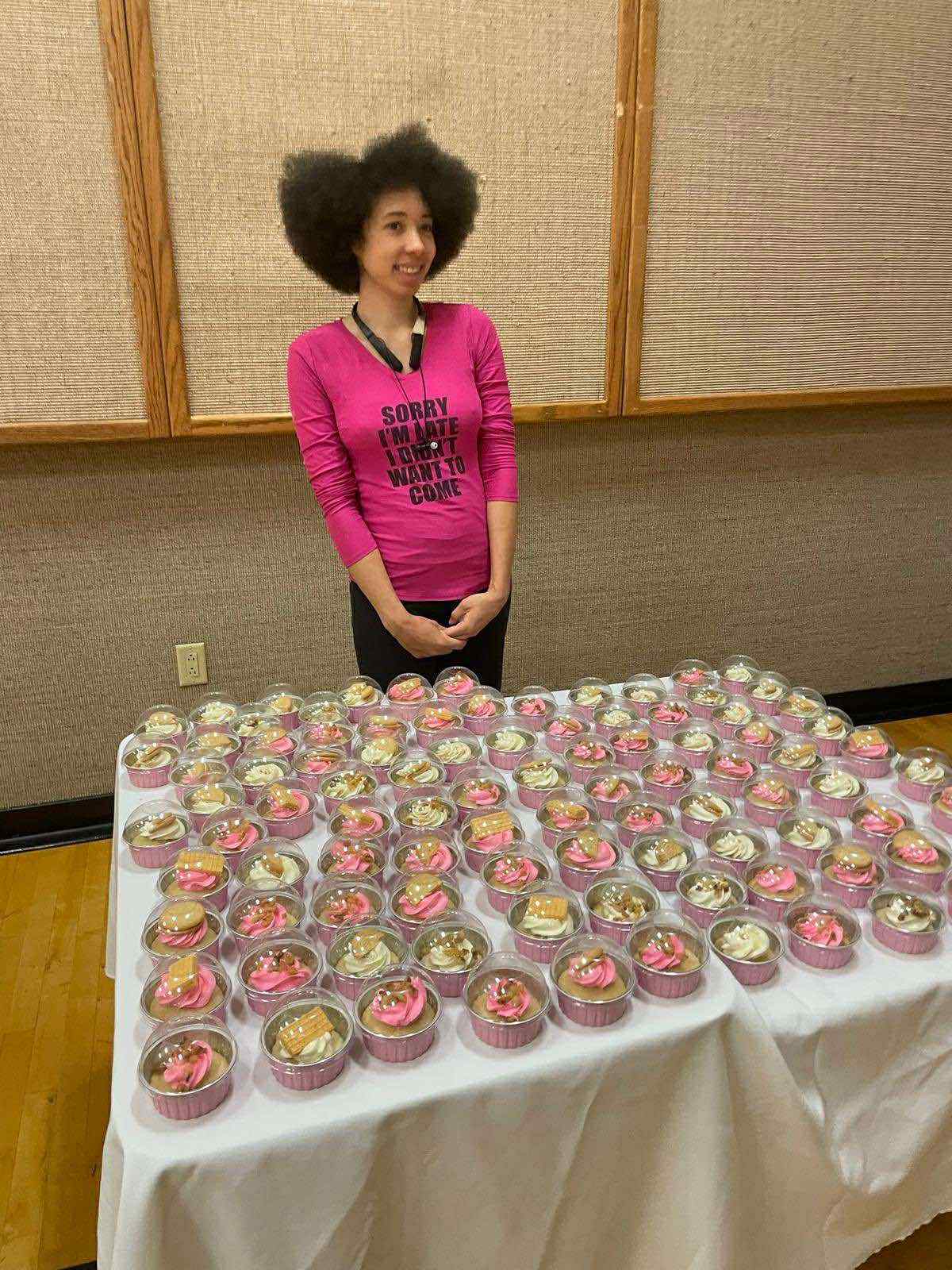 Young woman with curly hair standing behind a table filled with cupcakes, in a room with beige walls and wooden panels.