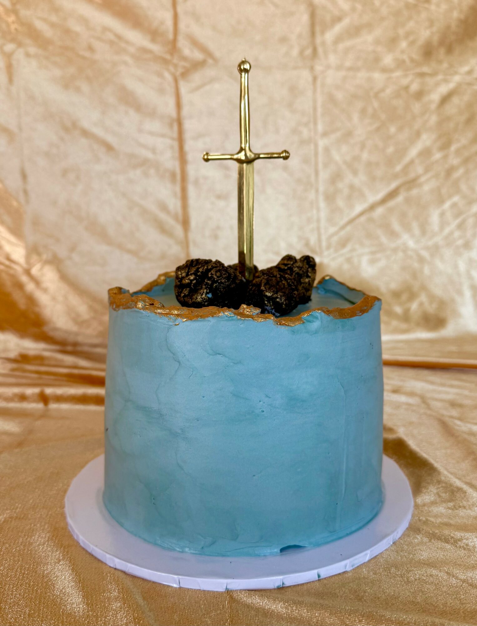A round cake with blue frosting, topped with a gold sword and black decorations, on a white base against a gold background.