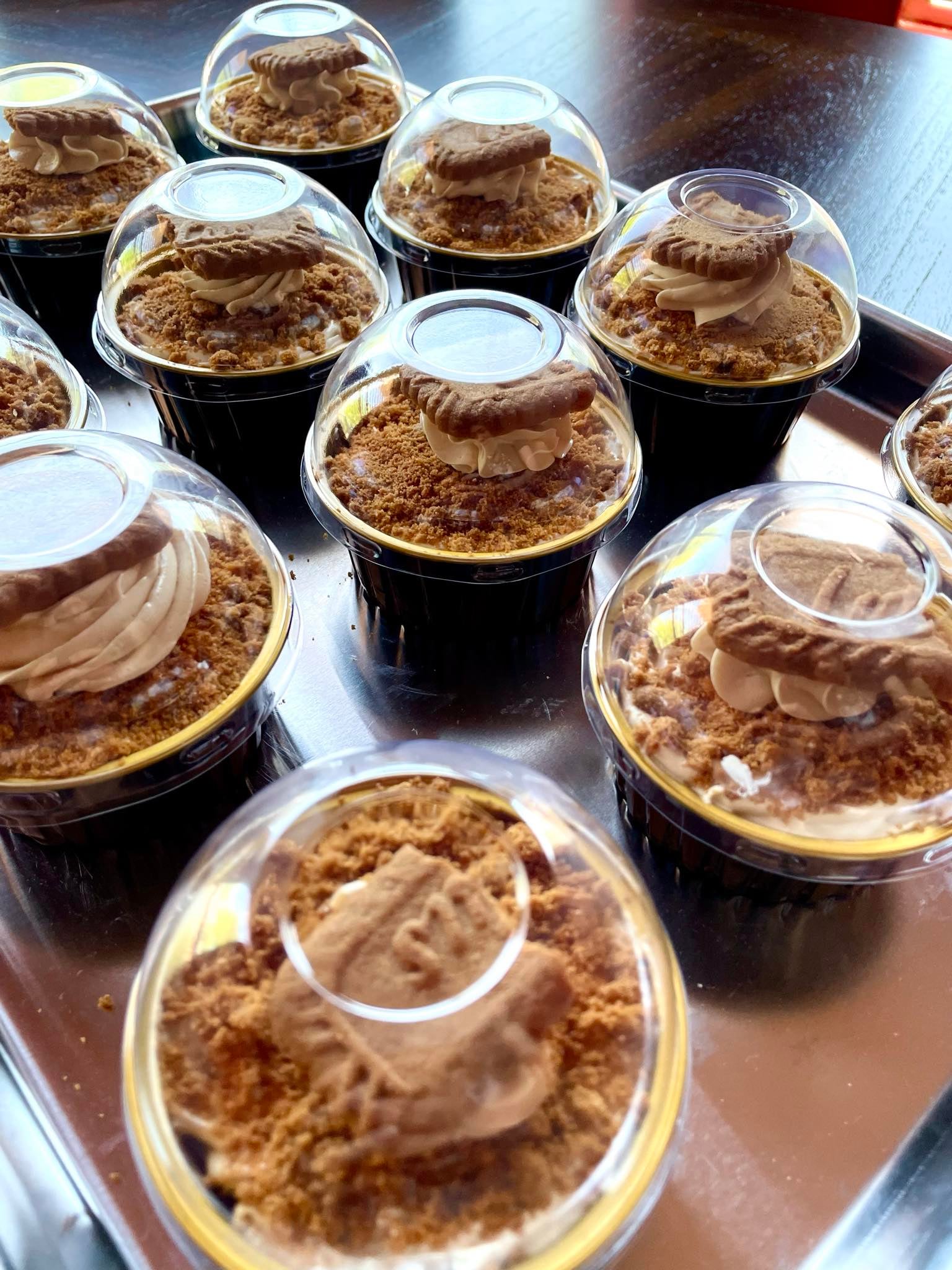 Multiple small desserts with layered cream and biscuit toppings in clear plastic cups on a table.