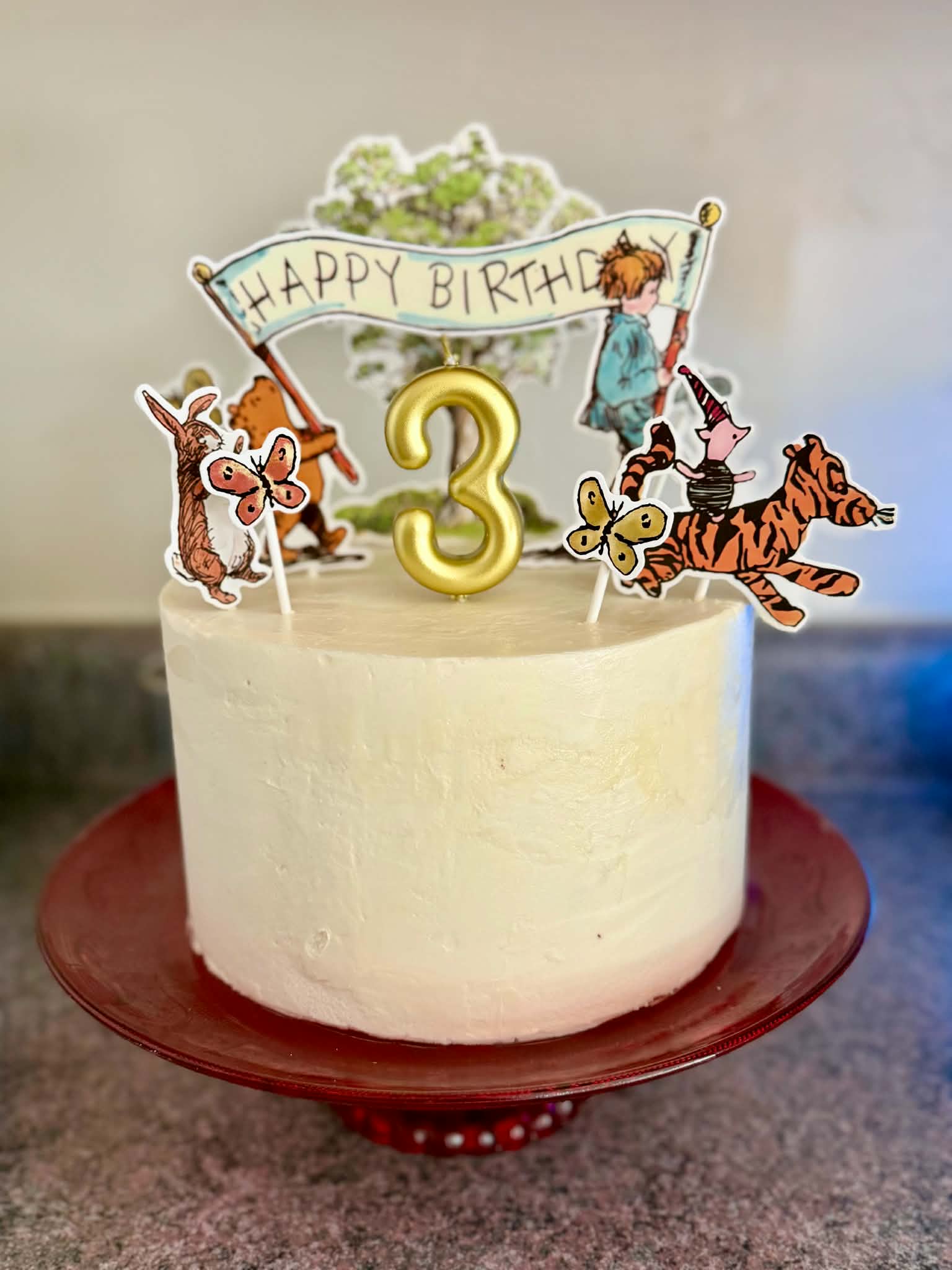 White birthday cake with animal and character decorations, a yellow number 3, and a banner reading 'Happy Birthday'.