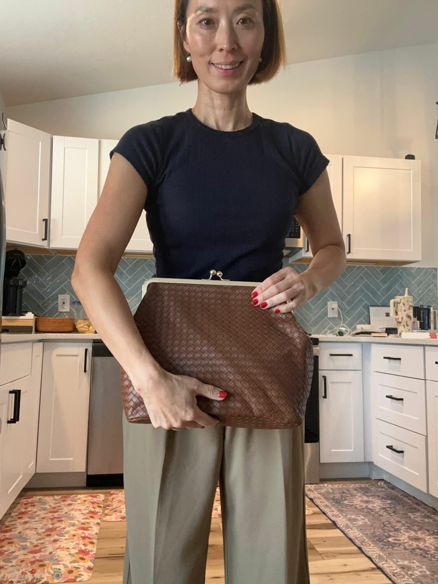 Woman standing in a kitchen holding a brown handbag, wearing a black top and beige pants, with kitchen cabinets and a rug in the background.