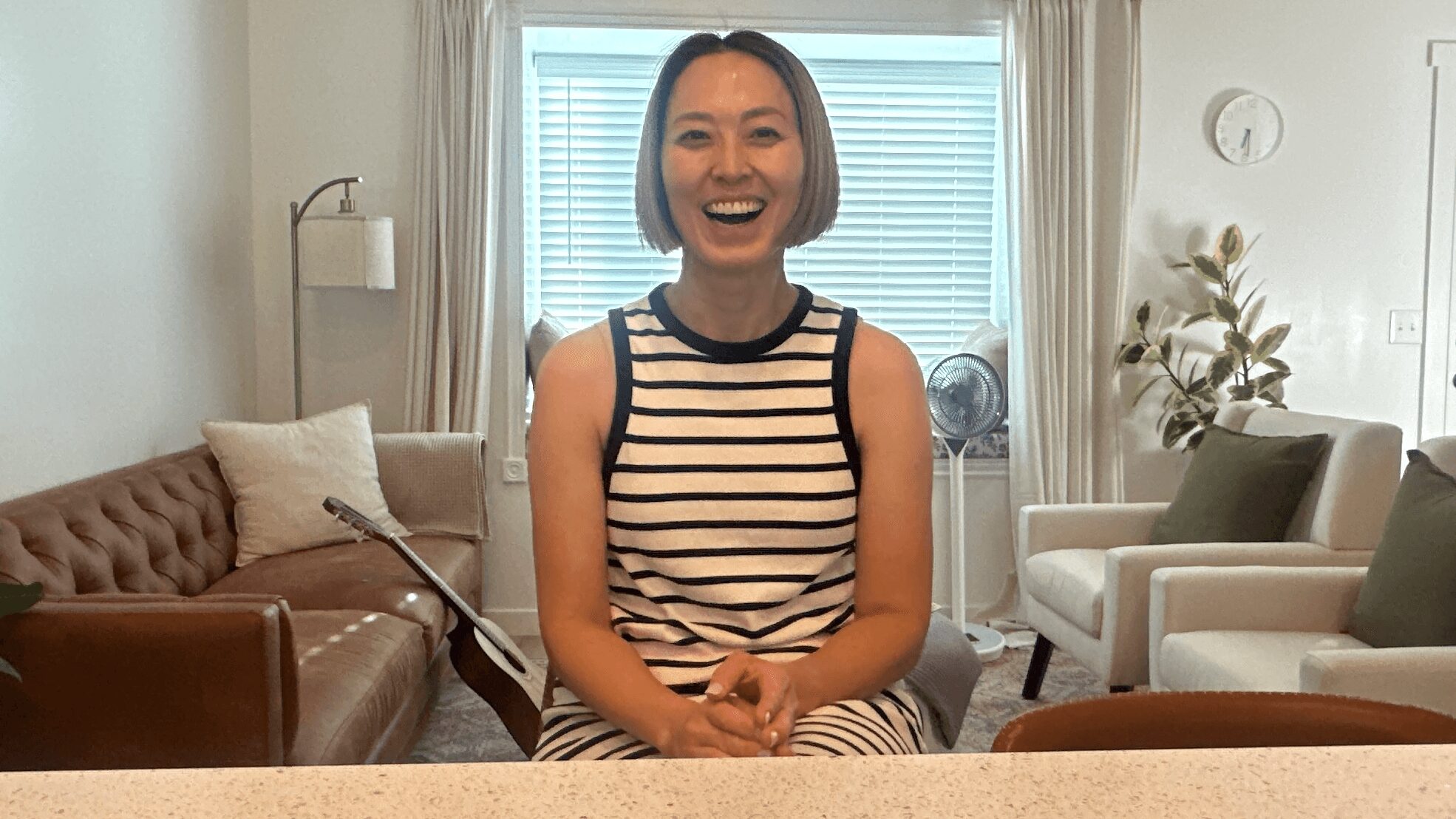 Smiling woman sitting at a table in a living room with furniture and plants behind her.