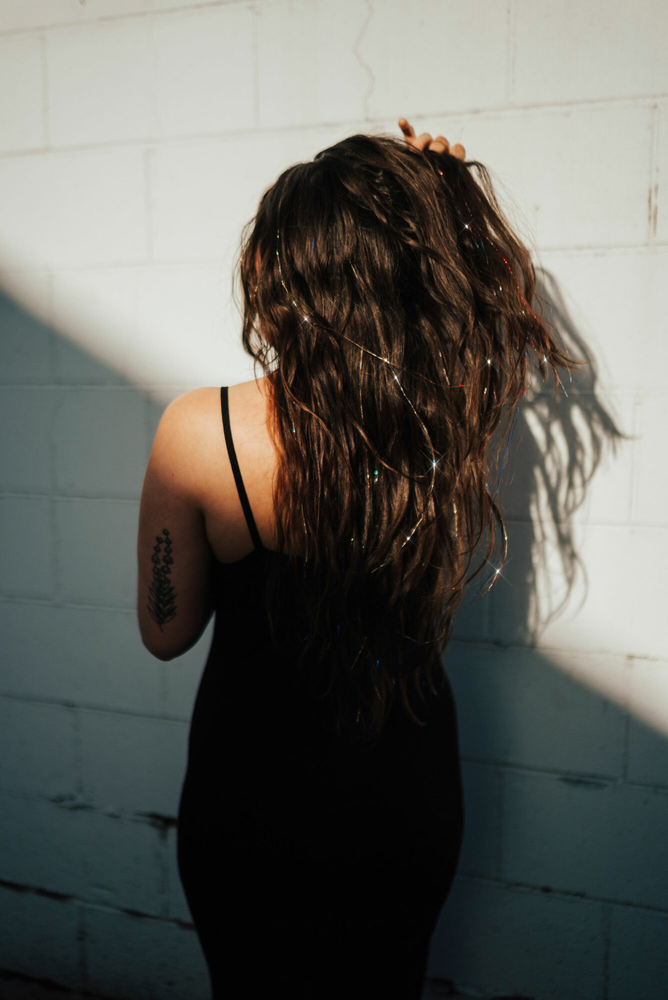 Woman with long, wavy hair wearing a black dress, standing against a white wall with shadow cast on it.