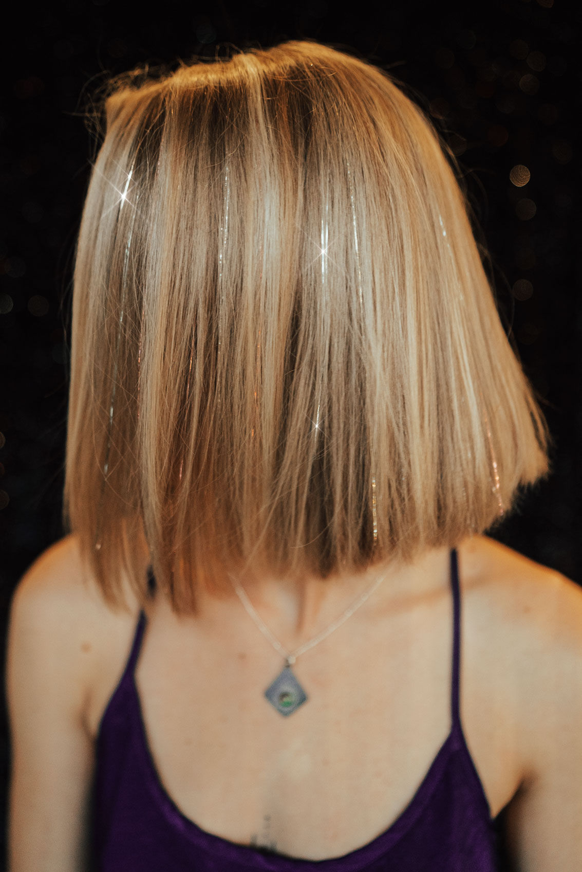 Person with shoulder-length blonde hair wearing a purple top and a necklace, facing away from the camera.