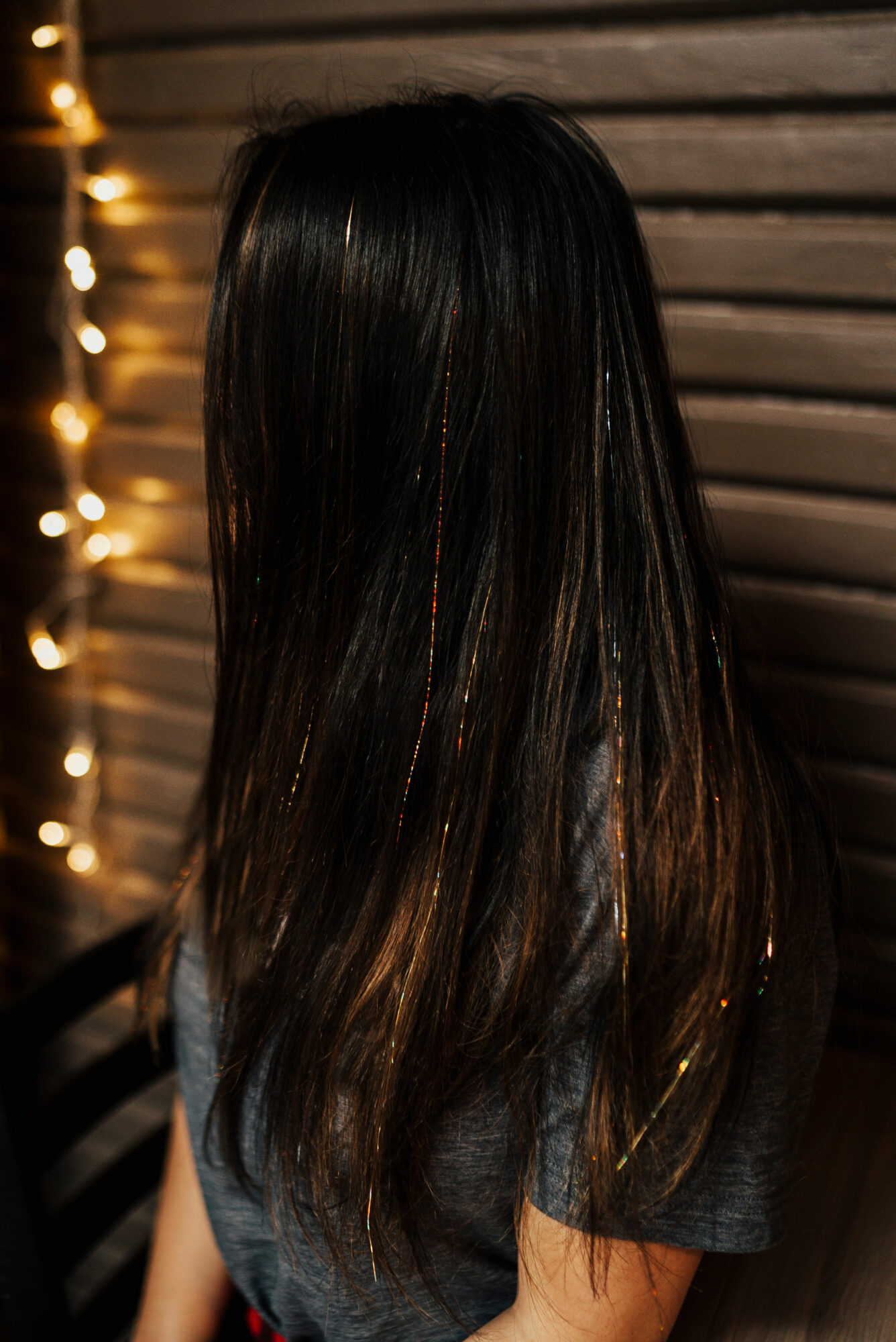 Back view of a person with long, dark hair standing against a wooden wall with string lights.