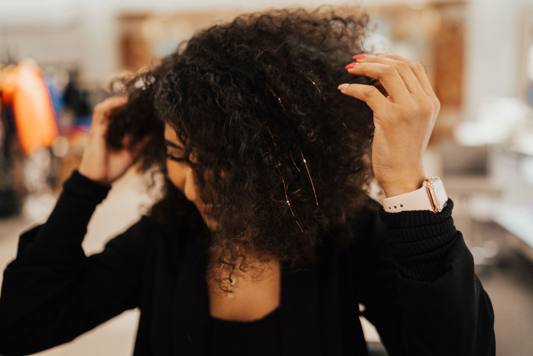 Person with curly hair adjusting hair in an indoor setting, blurred background, wearing a black top and a watch.