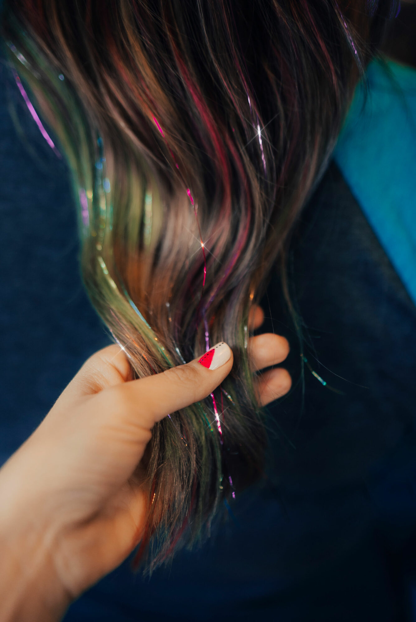 Hand holding long, wavy, multicolored hair with pink, purple, and blonde streaks, against a dark background.