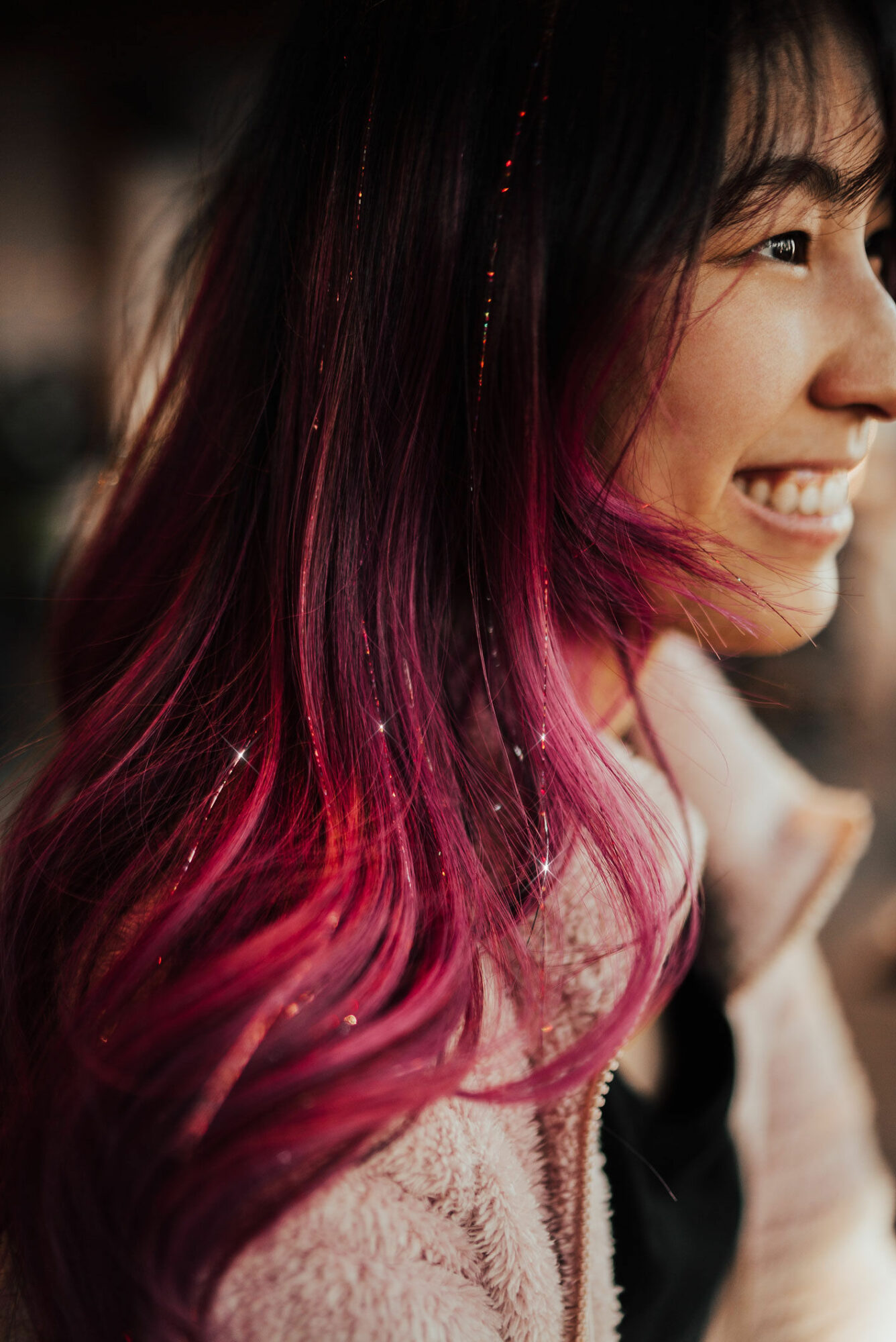 Close-up of a smiling woman with long, dark hair with pink tips, wearing a light-colored coat, outdoors.