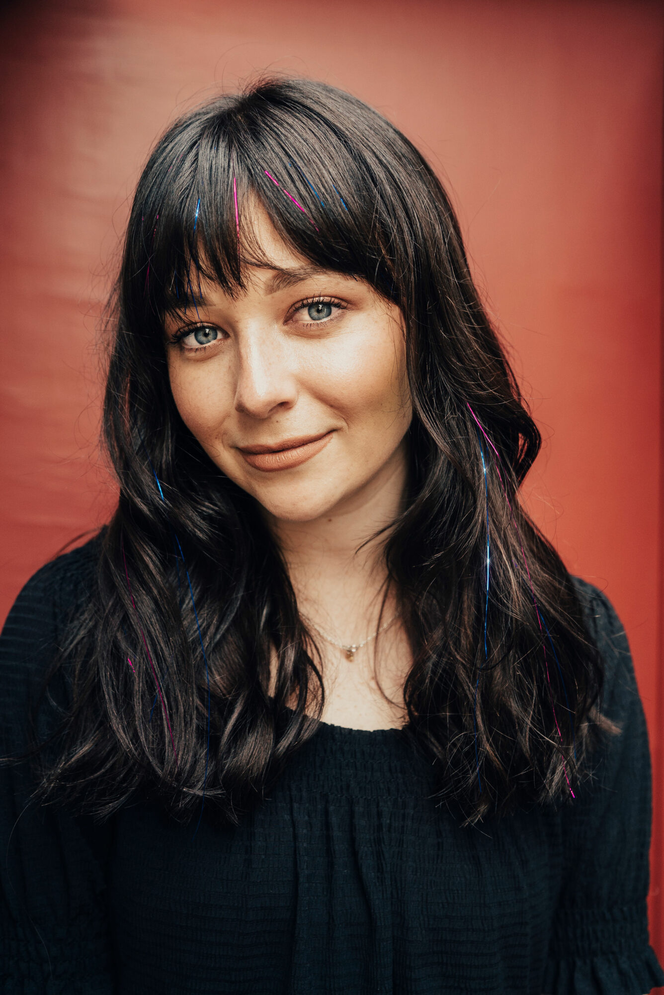 Young woman with dark wavy hair and bangs, smiling, wearing a black top, against a reddish background.