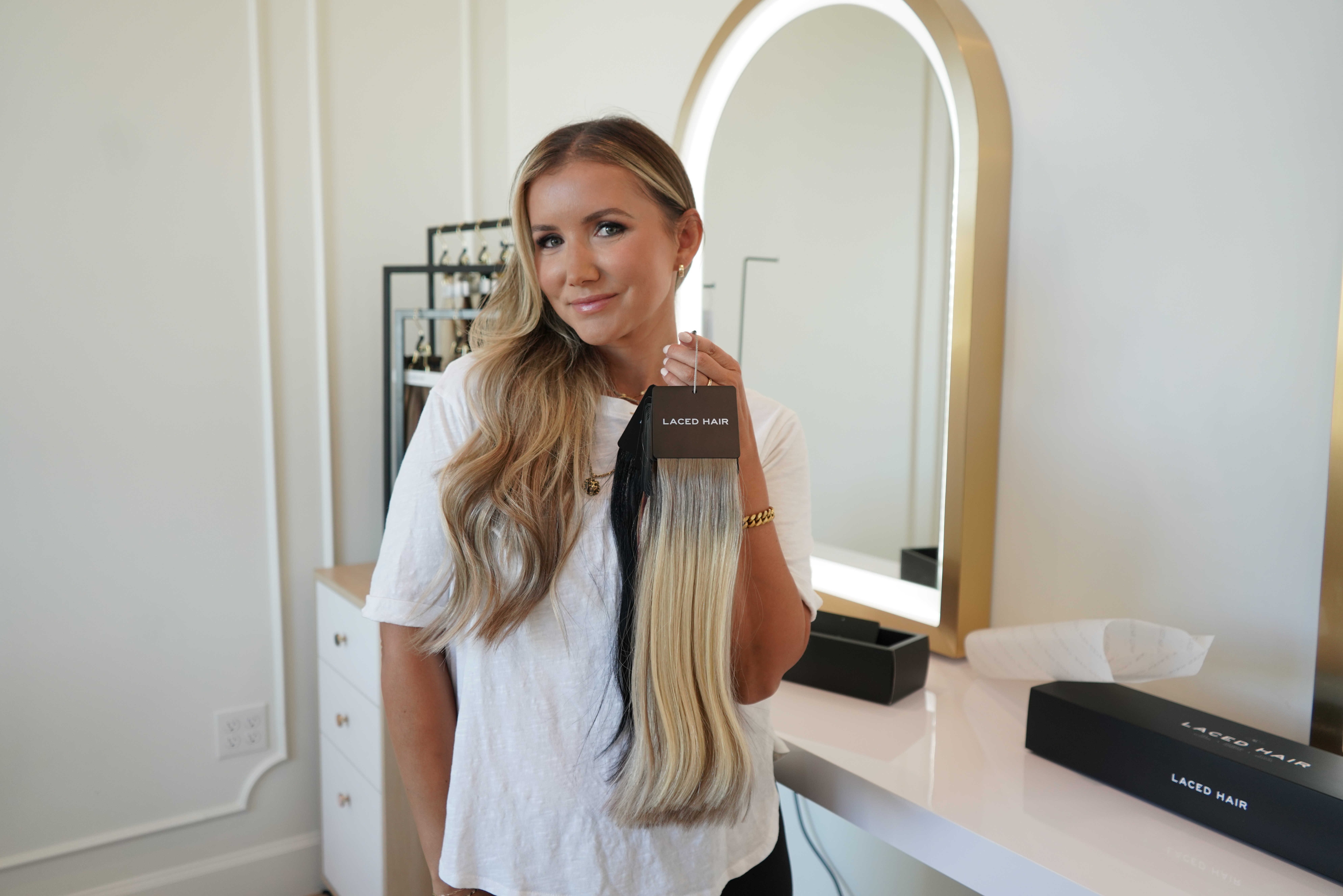 Woman with long blonde hair taking a mirror selfie in a bright room, smiling, wearing a white top.