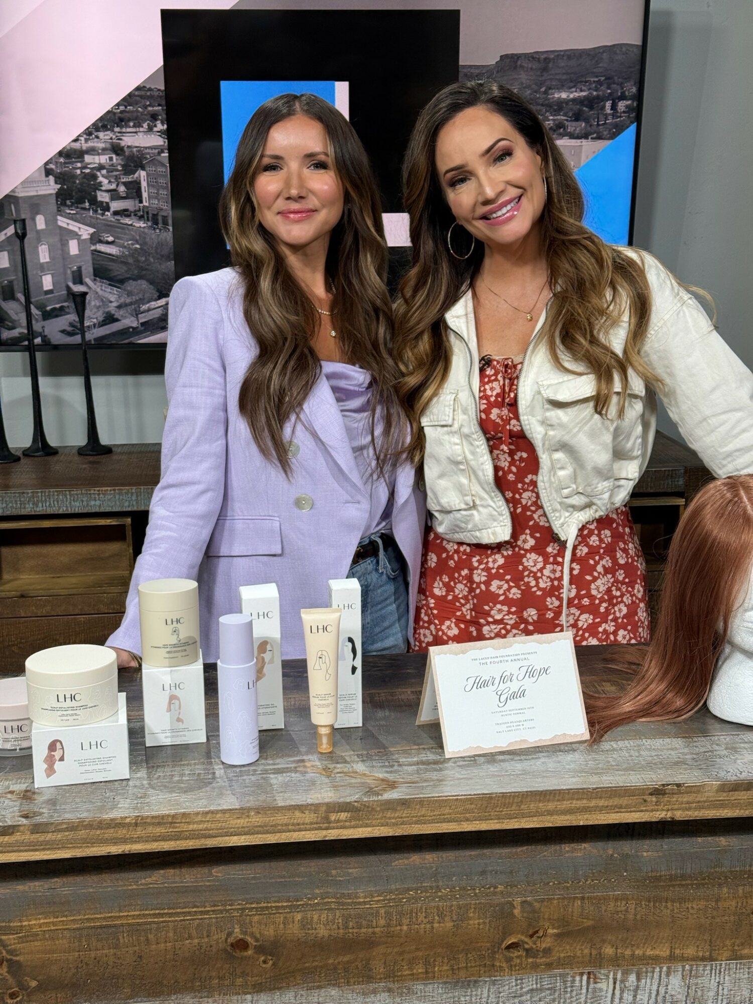Two women stand behind a table with skincare products, smiling, in an indoor setting.