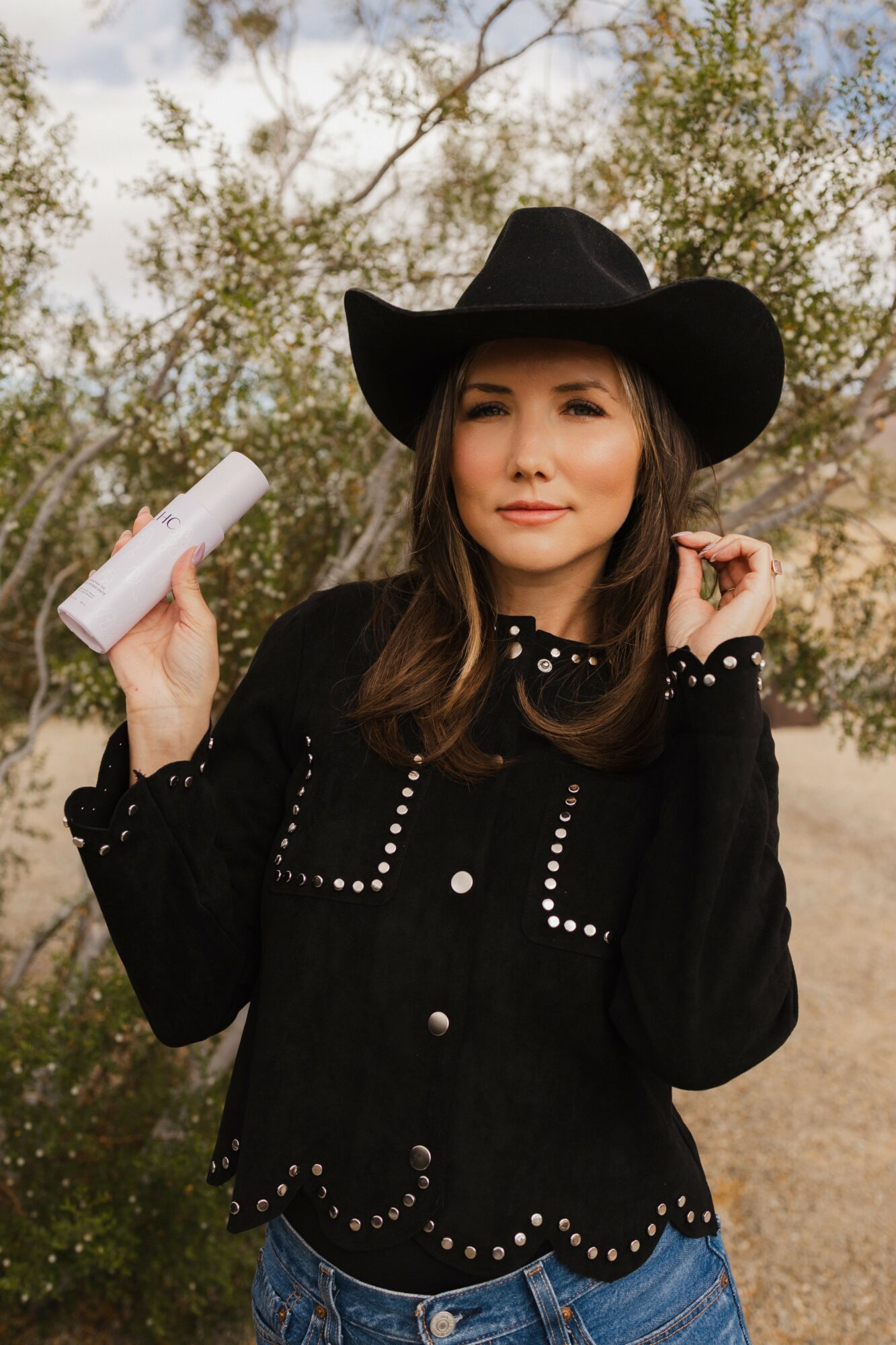 Woman wearing a black hat and jacket, holding a white bottle outdoors with trees in the background.