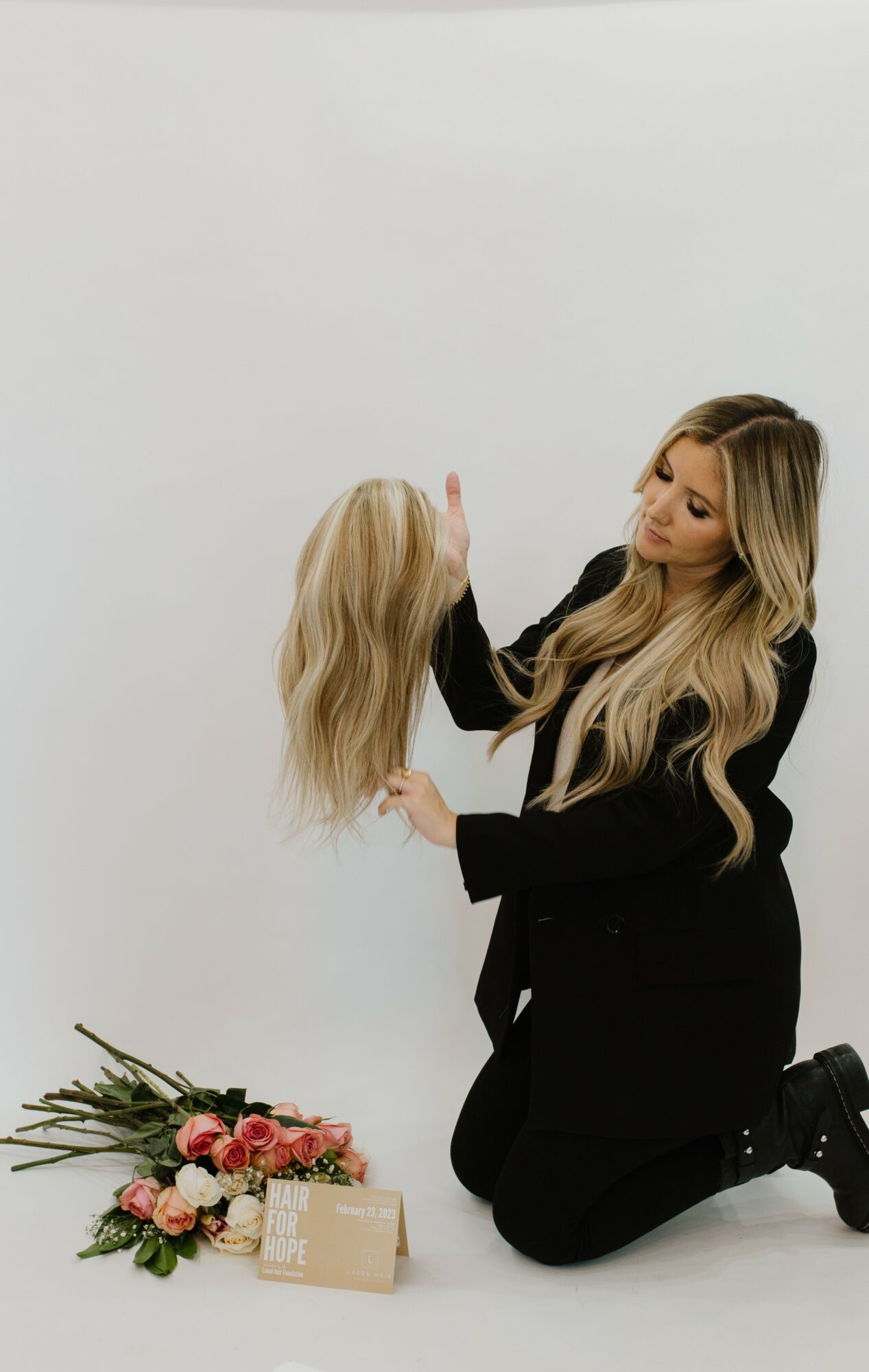 Woman kneeling on the floor holding a blonde wig, with flowers and a gift box nearby, against a plain white background.