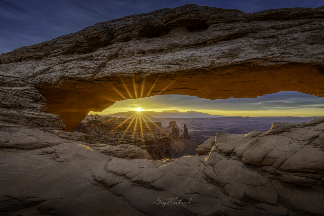 Sunset viewed through a natural rock arch with a clear sky and distant landscape, warm colors, and sun rays.