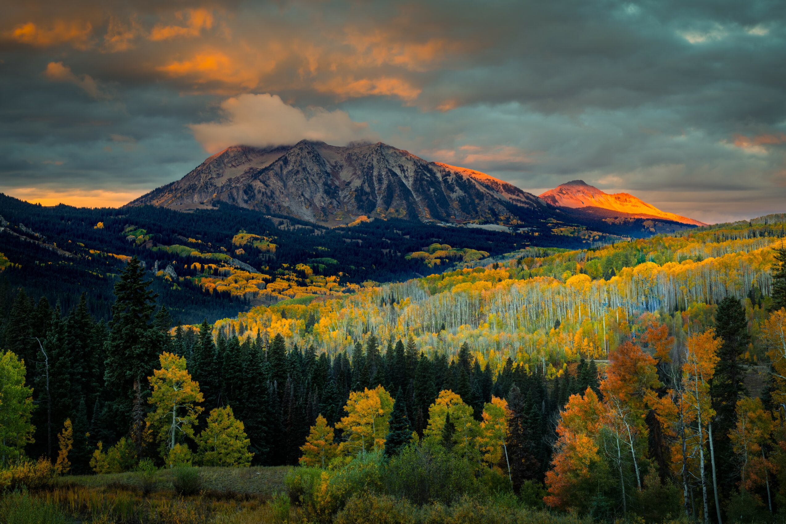 Mountain with snow and clouds above a forest with colorful autumn trees, sunset lighting, and a cloudy sky.