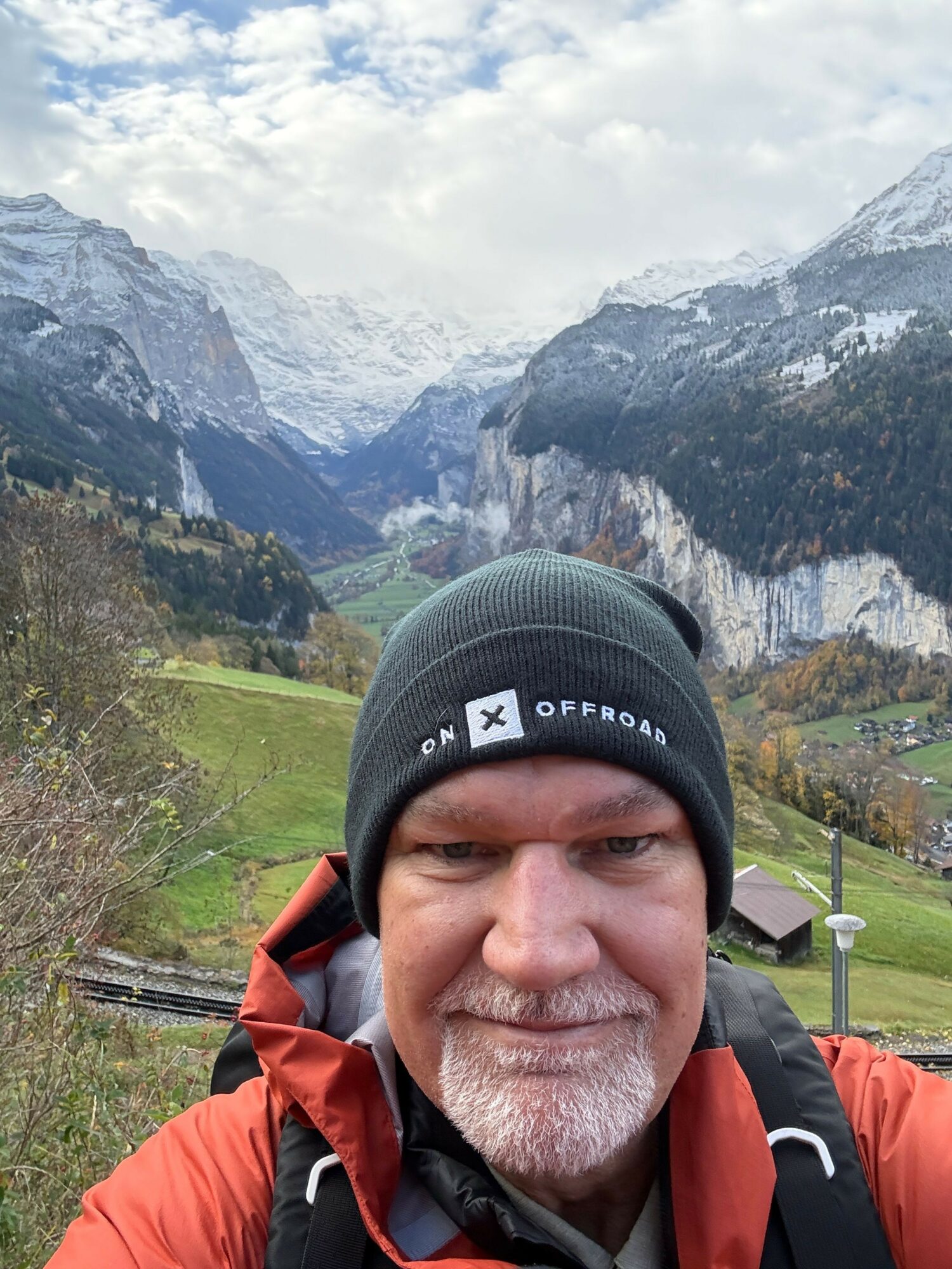 Man outdoors with mountains, cloudy sky, and green fields in background, wearing a hat and outdoor gear.