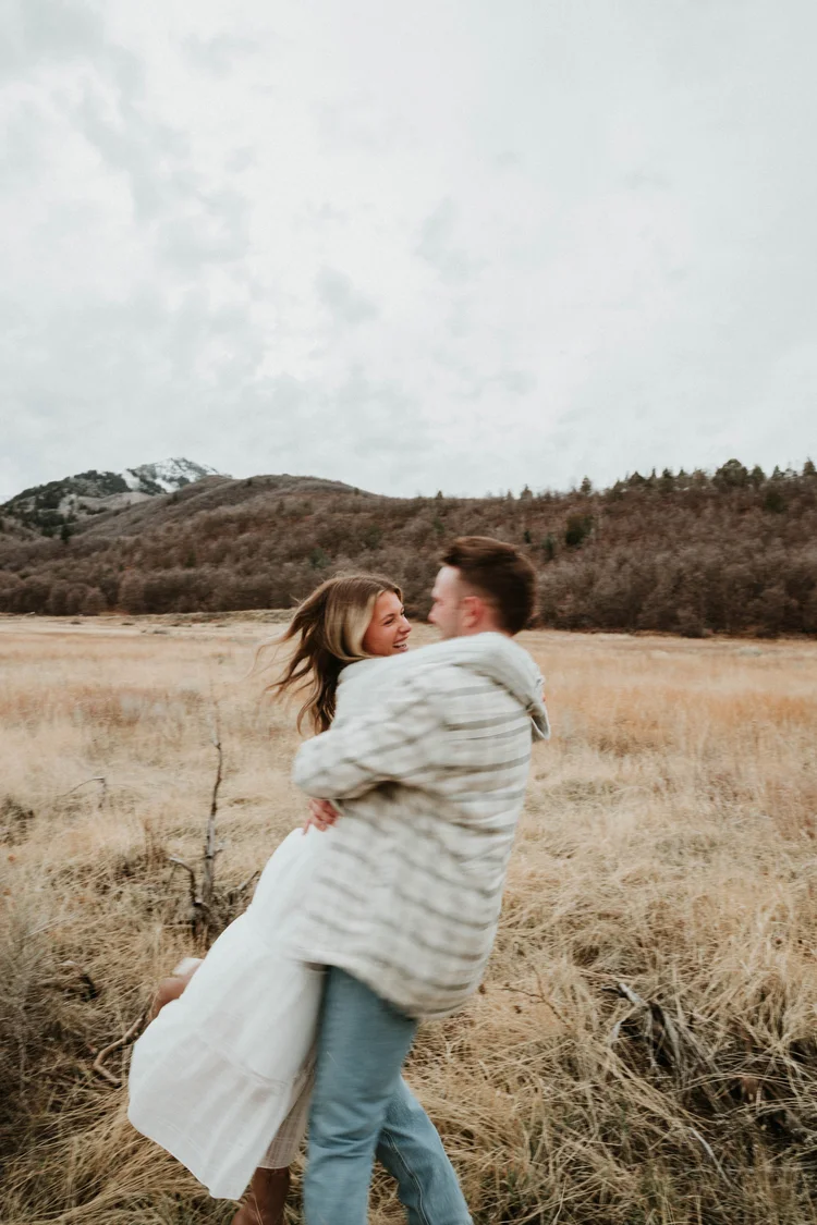 A couple embracing in a field with mountains and cloudy sky in the background.