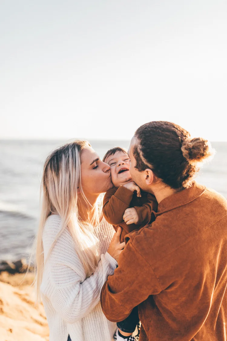 Two women kiss a smiling child at the beach during sunset, with ocean and sky in background.