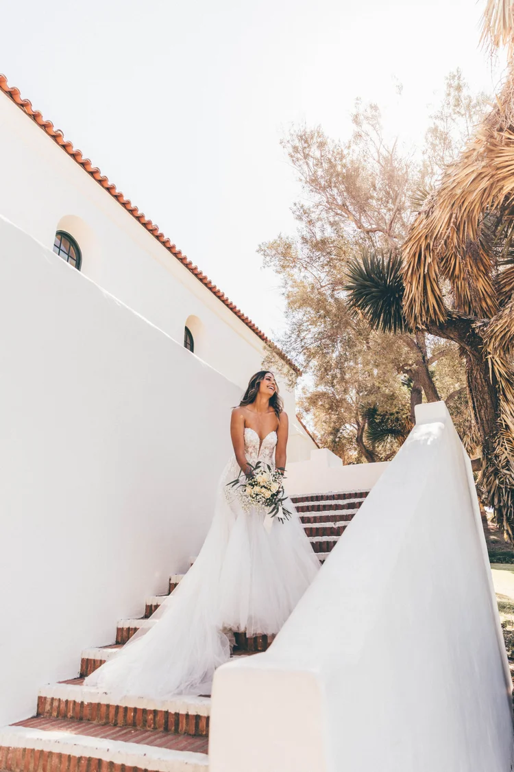 Bride in a white wedding dress holding a bouquet, standing on stairs outside with trees and a white wall behind.