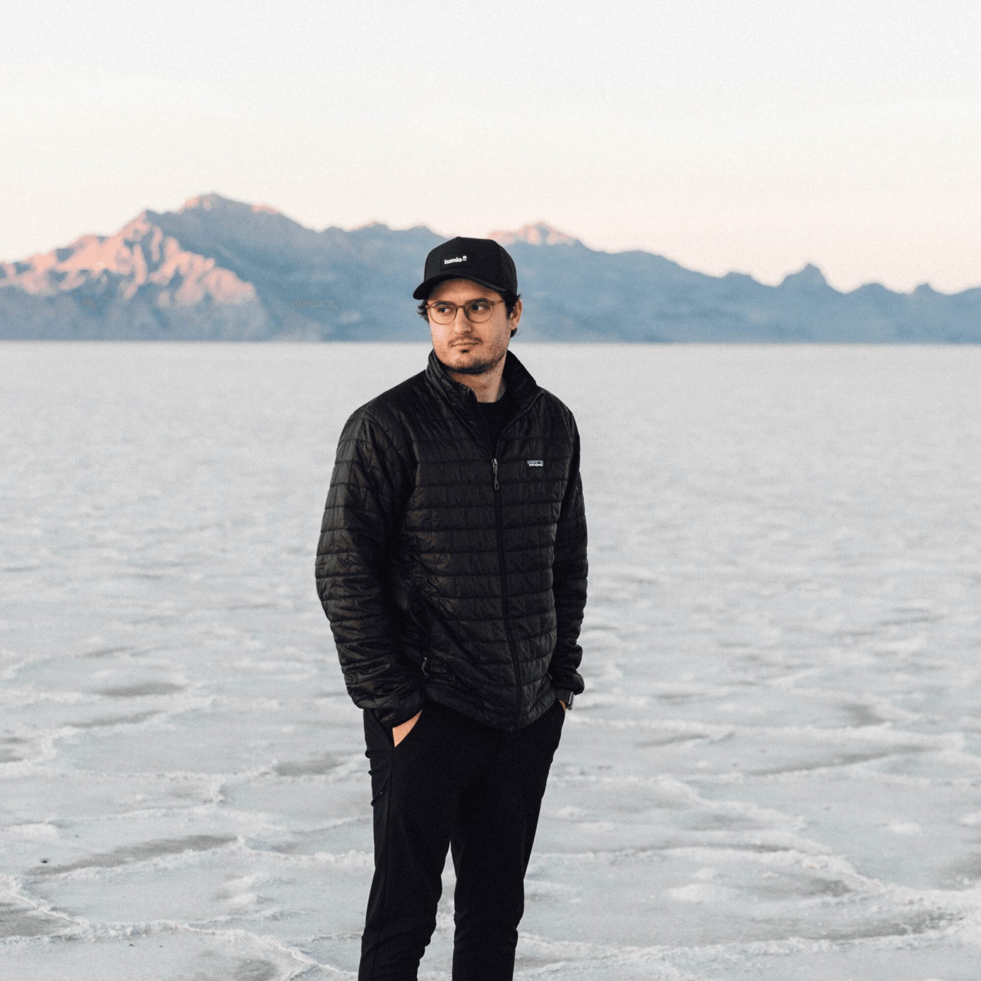 Man standing outdoors near water with mountains in background, wearing glasses, a black cap, and a black jacket.