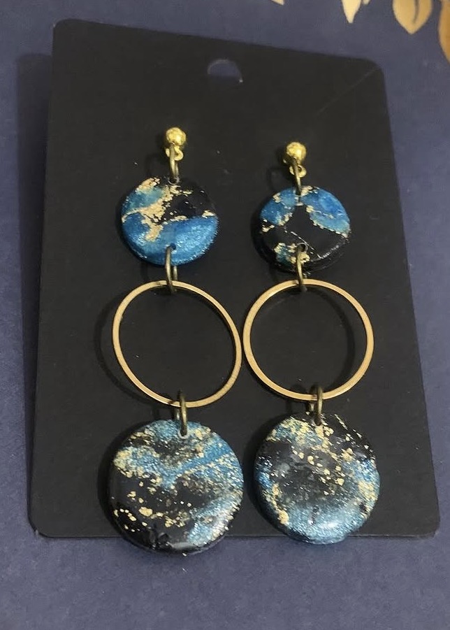 Pair of earrings with circular blue and black designs, gold rings, and gold accents, displayed on black background.