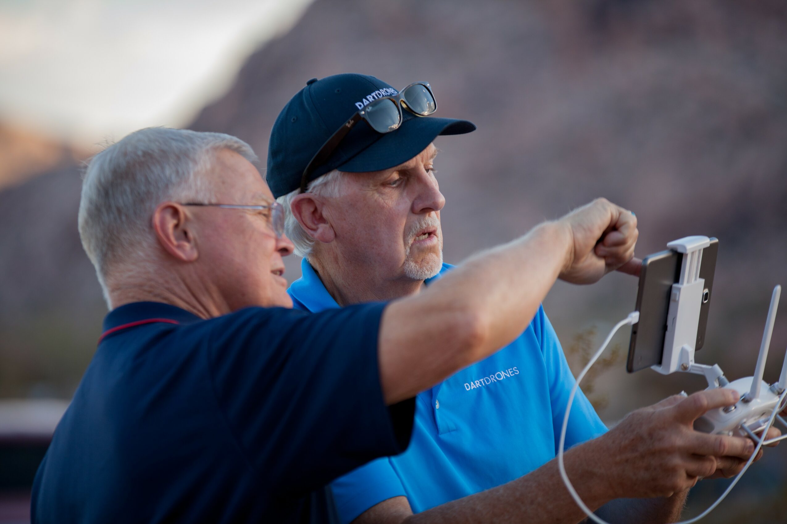 Two men look at a drone controller outdoors, one pointing at the device, with a blurred natural background.