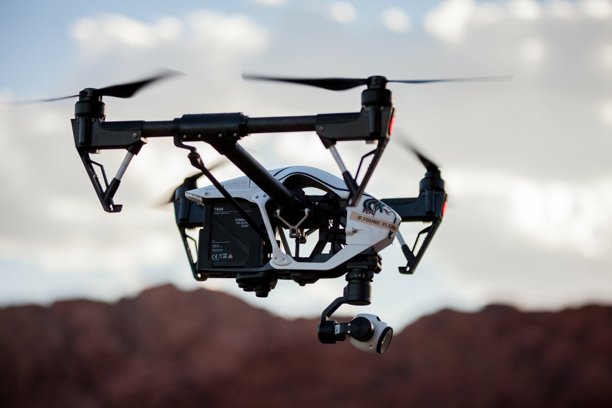 Quadcopter drone flying outdoors with a camera attached, sky and landscape in background.