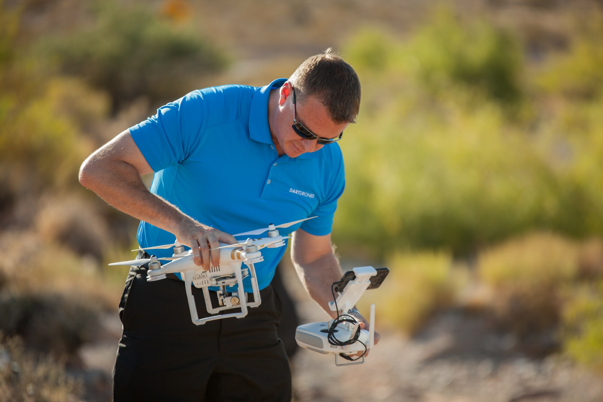 Man in blue shirt holding a drone controller outdoors with a desert landscape background.