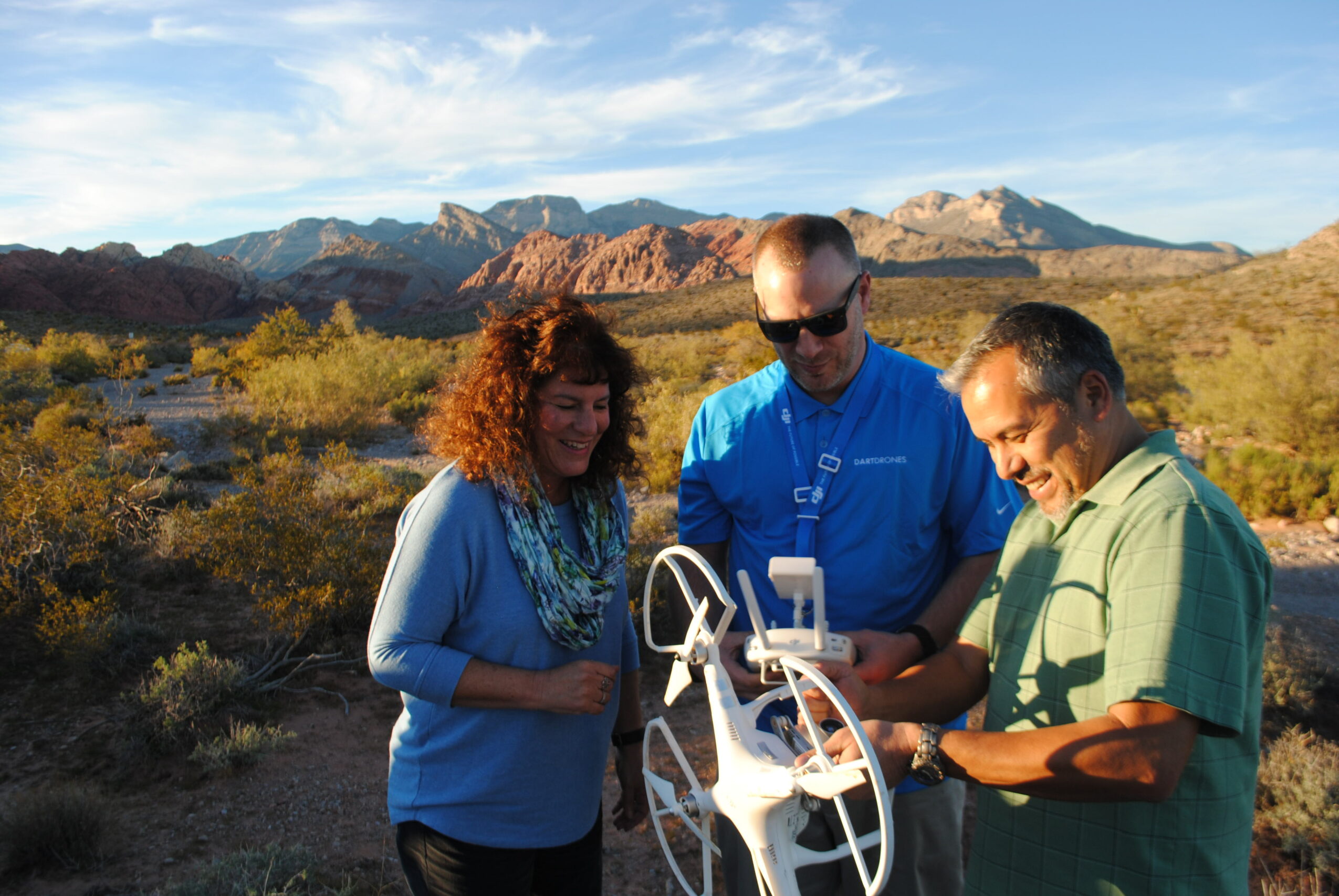 Three people outdoors in a desert landscape with mountains, looking at a device, smiling, during daytime.
