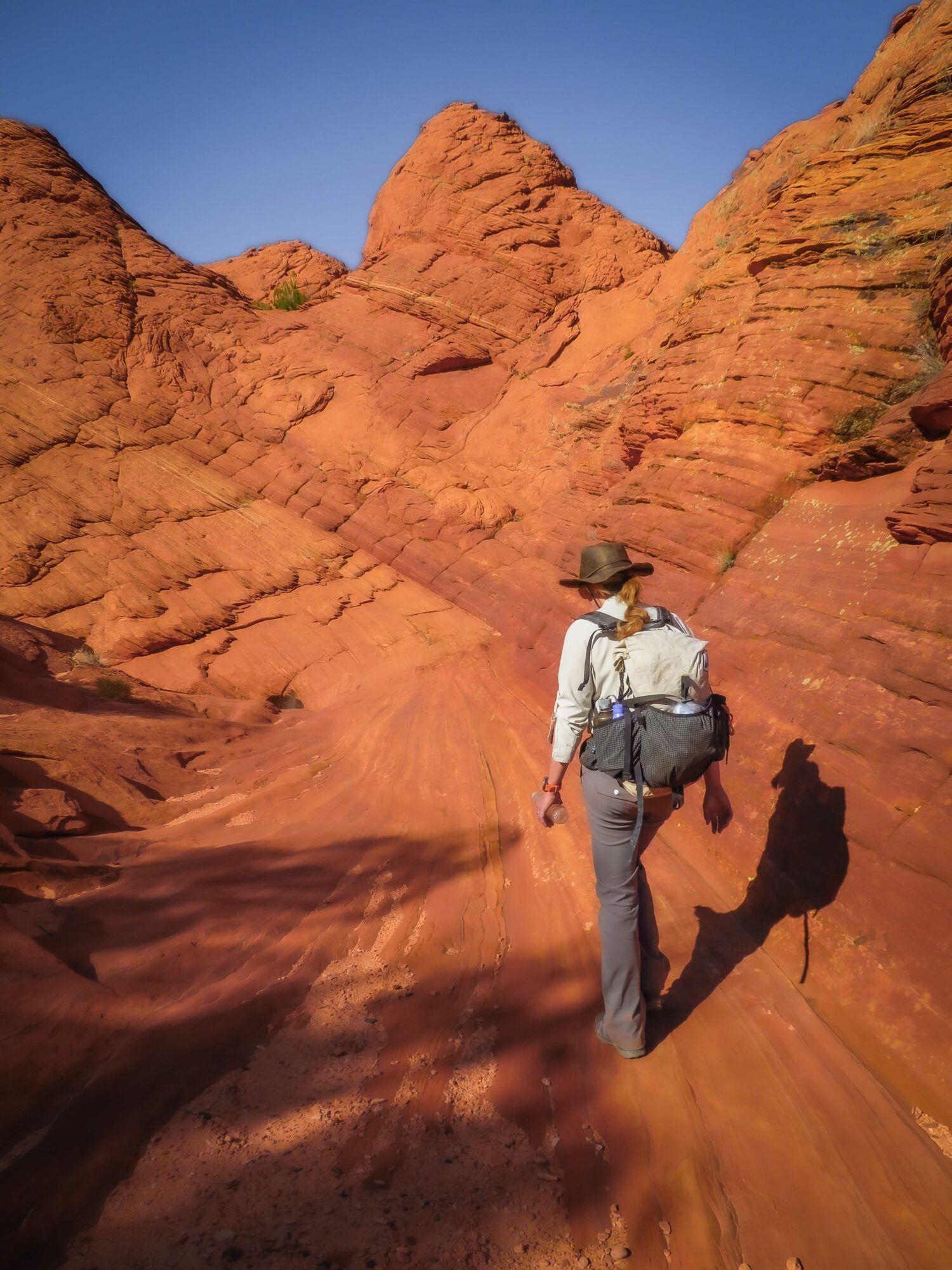 Person walking in a red rocky desert landscape with clear blue sky, carrying a backpack and wearing a hat.