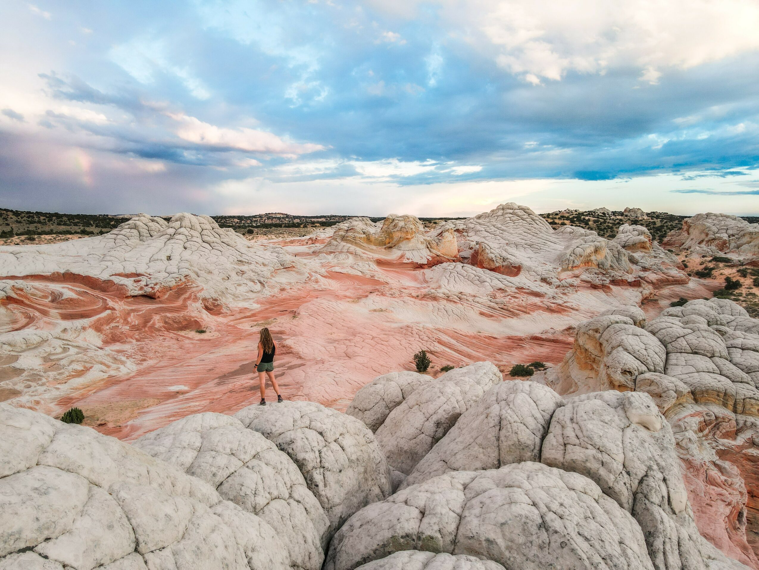 Person standing on colorful layered rock formations with a cloudy sky above, in a desert landscape.