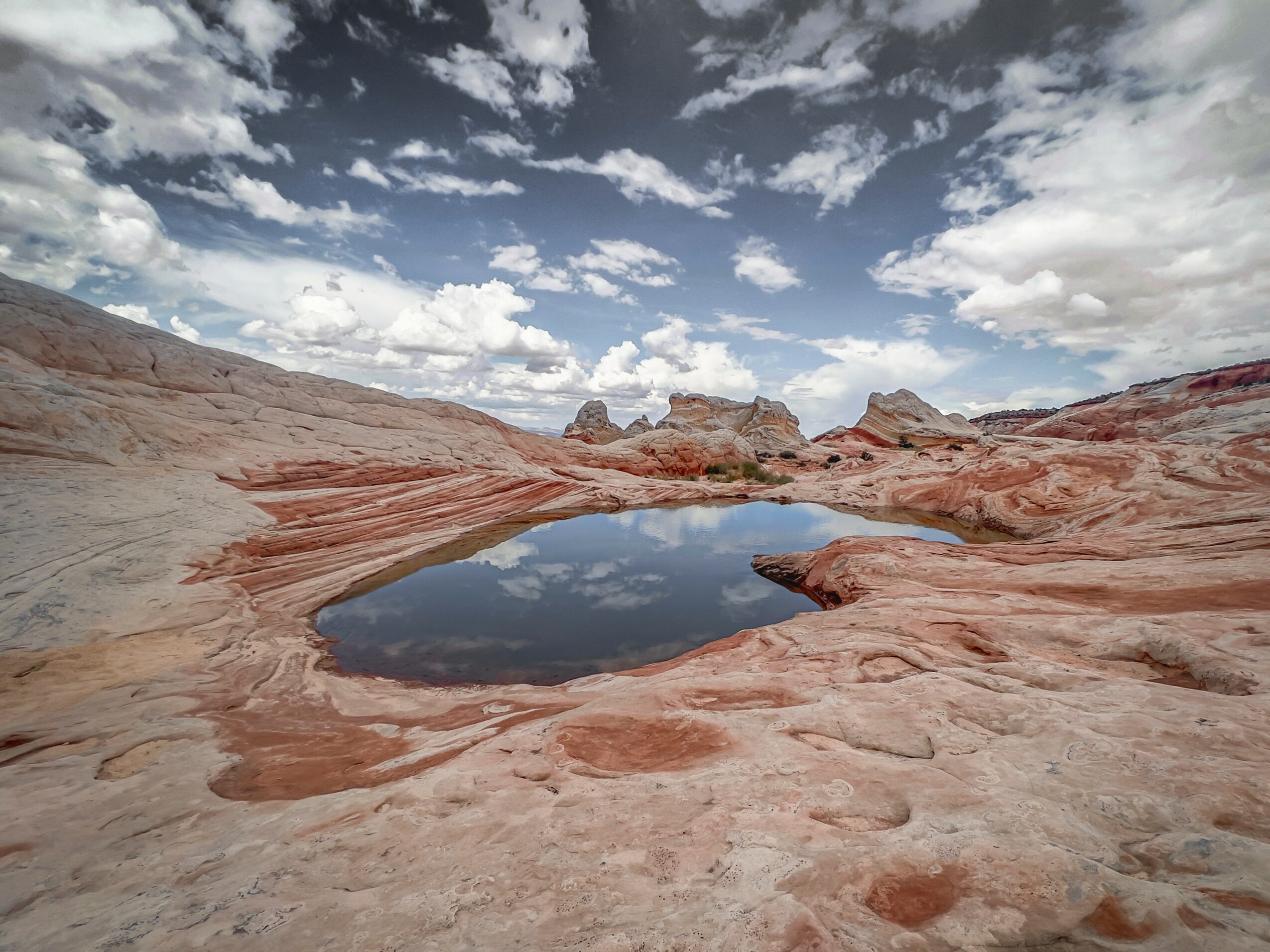 A small pond reflects the sky with clouds, surrounded by reddish rock formations in a desert landscape.