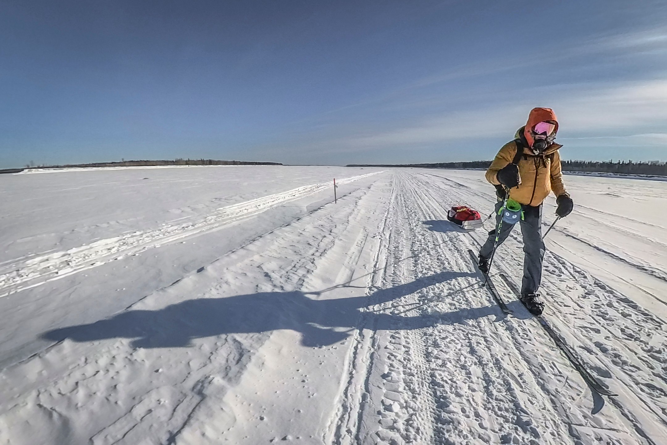 Person in winter clothing with a backpack and ski poles walking on snow-covered landscape under a clear sky.