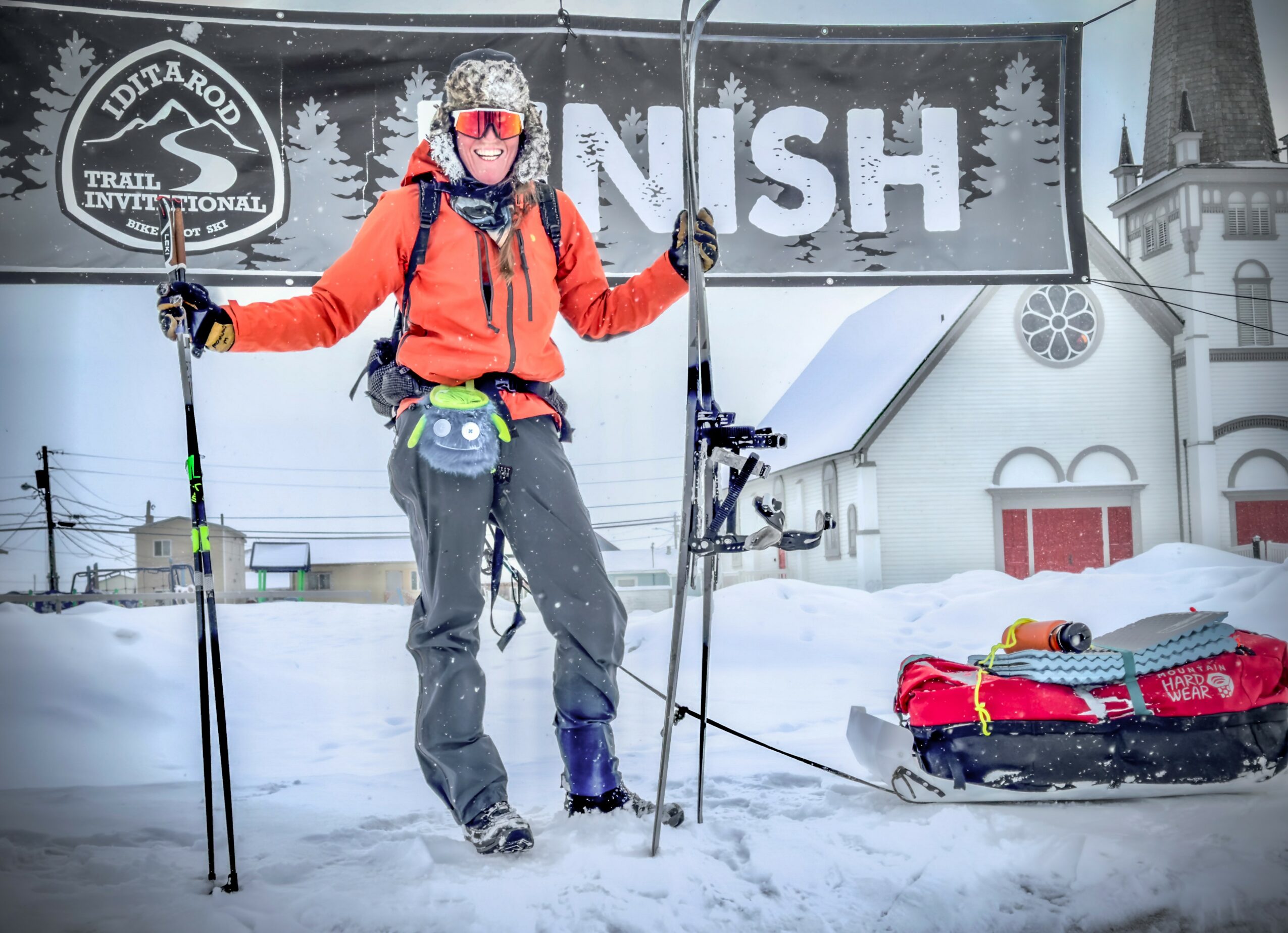 Person in winter gear with ski poles and a sled in snowy landscape, standing in front of a building with trees and a sign.