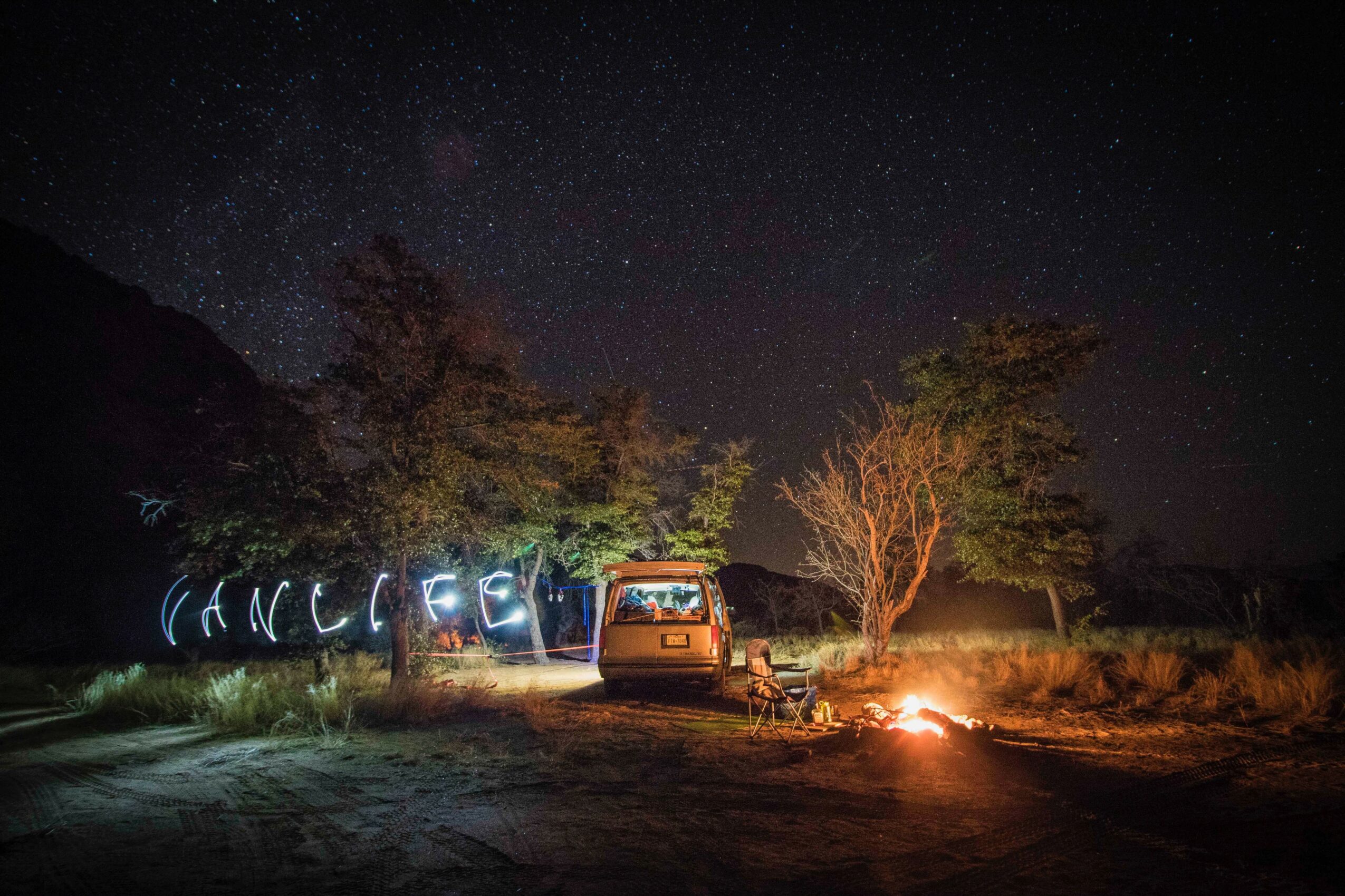 Night scene with a van, trees, and a campfire under a starry sky, with light trails spelling 'CANCELLED'.