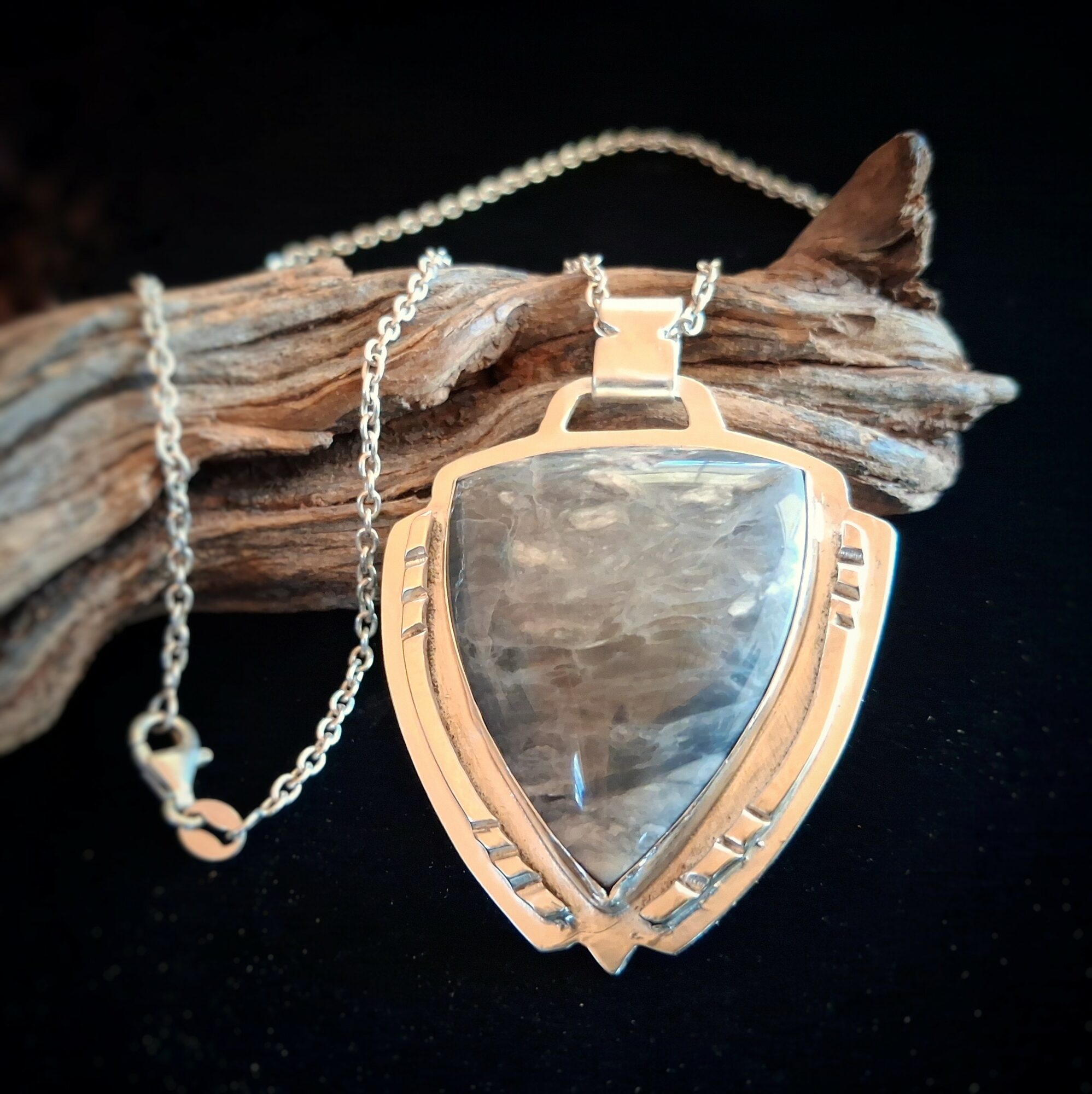 Pendant with a large, teardrop-shaped stone in a gold setting, attached to a chain, resting on a piece of driftwood.