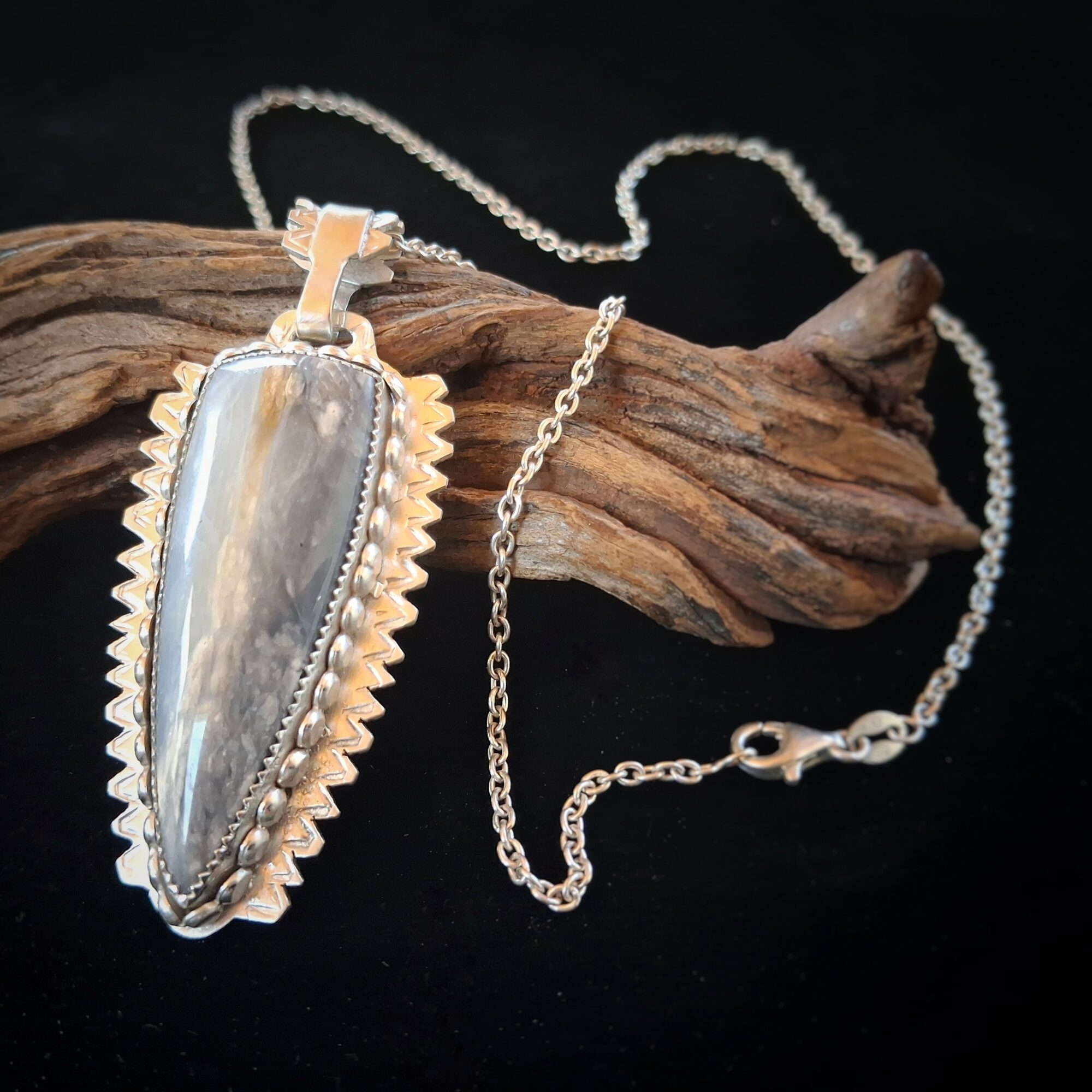 Pendant necklace with a large, elongated, iridescent stone on a silver chain, resting on a piece of wood against a black background.