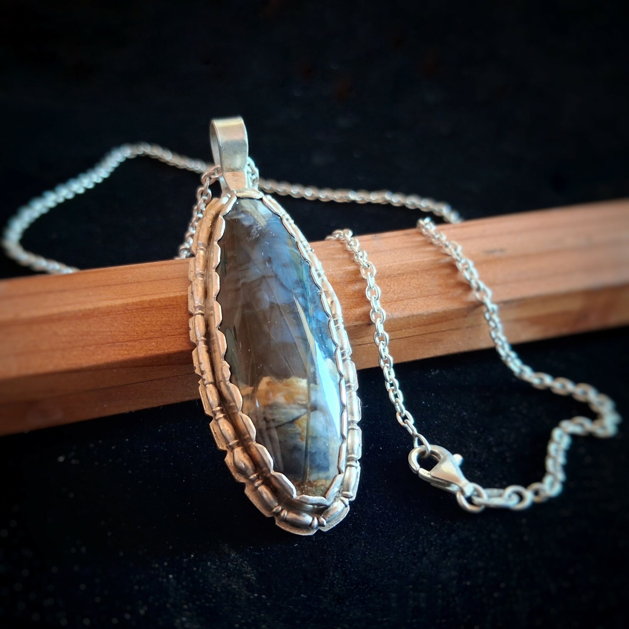 Pendant necklace with a large oval gemstone on a silver chain, resting on a wooden surface, against a dark background.