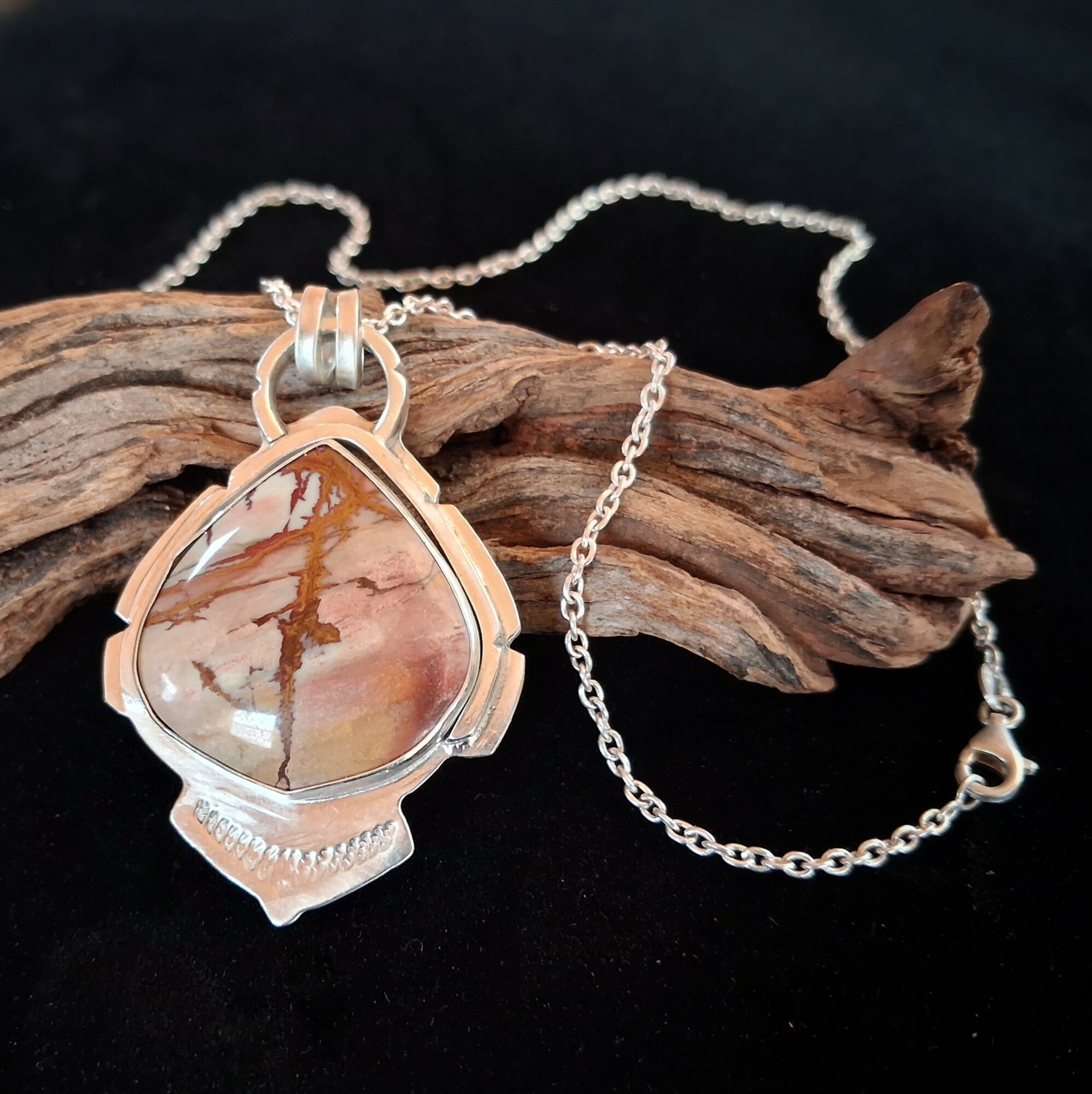 Silver necklace with a large, oval-shaped pendant featuring a pink and brown marbled stone, resting on a piece of driftwood.