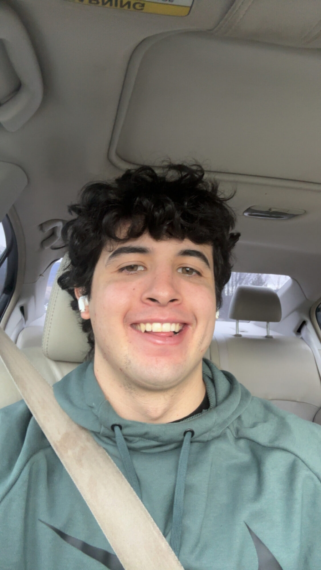 Young man with curly dark hair smiling inside a car, wearing a green hoodie and seatbelt.