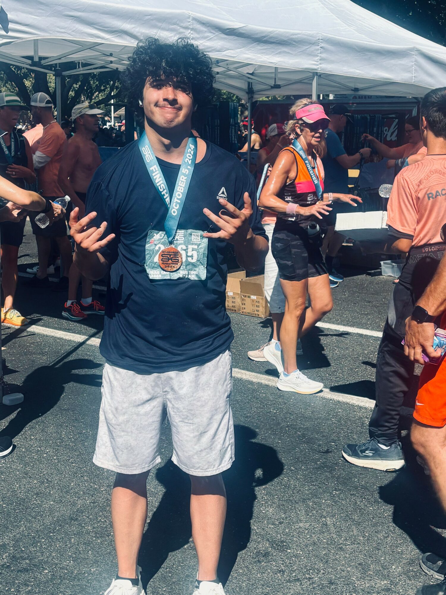 Young man with curly hair wearing a medal, black shirt, and light shorts, standing outdoors among people at an event.