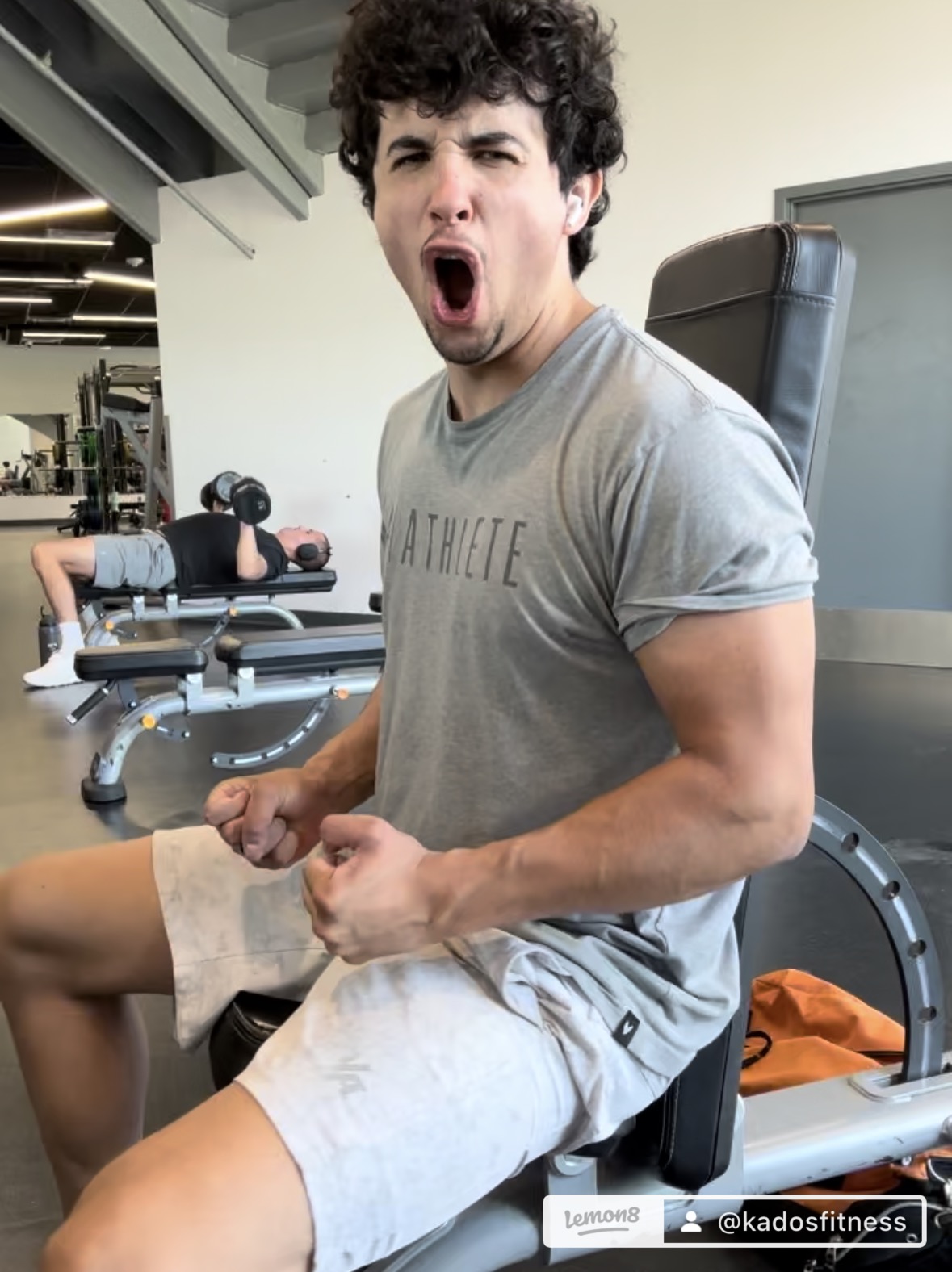Young man at gym flexing arm muscles with mouth open, wearing gray t-shirt and shorts, gym equipment in background.
