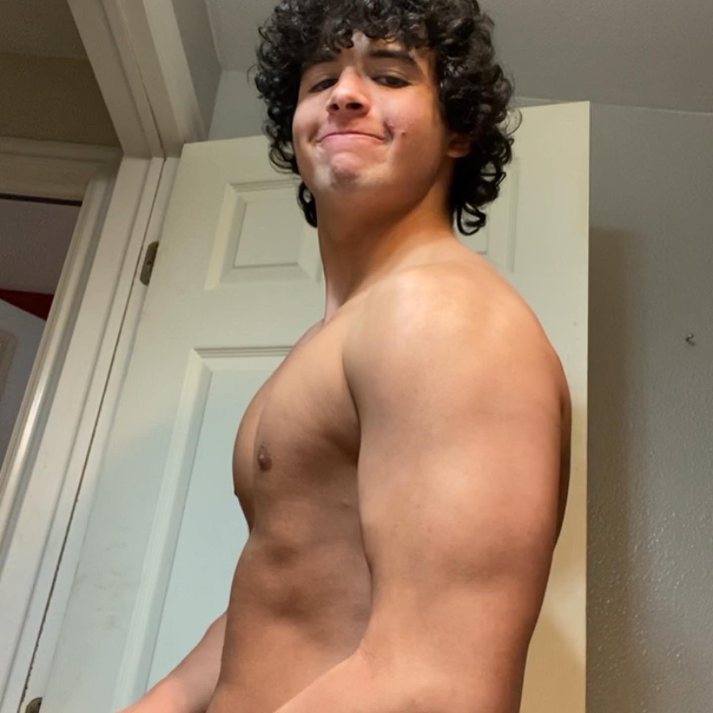 Young person with curly hair smiling, shirtless, standing indoors near a door, facing slightly to the right.