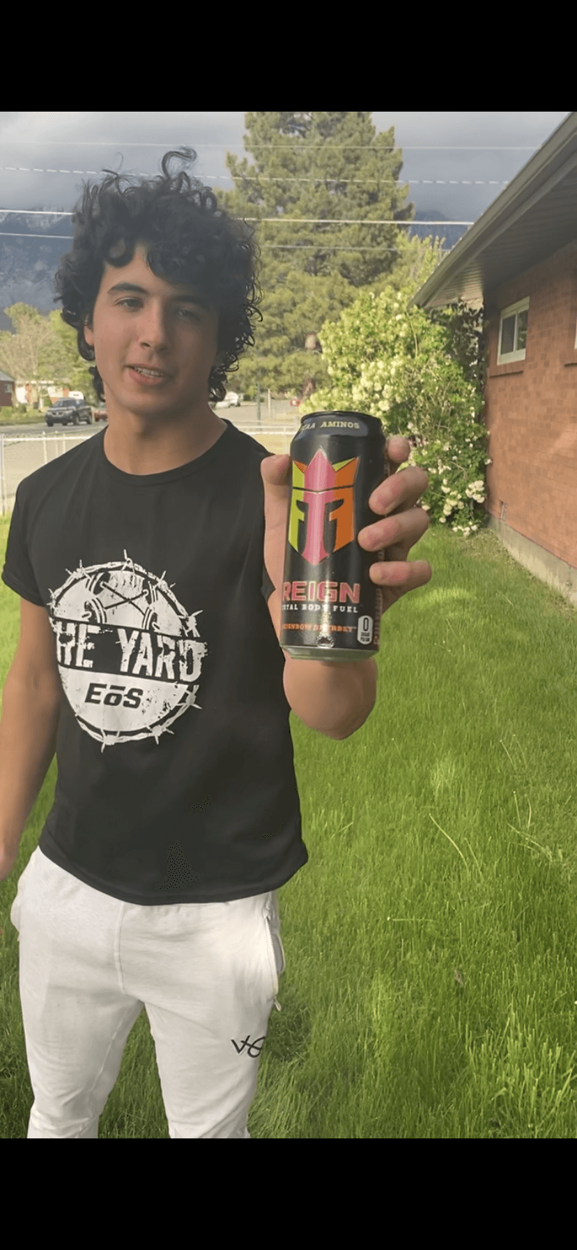 Young person with curly hair holding a can outdoors, standing on grass with trees and a building in background.